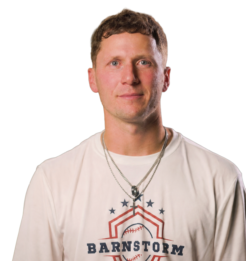 A young man with short brown hair and blue eyes is smiling slightly. He is wearing a white sports jersey with a baseball and star design and the word 'Barnstorm' printed on it. He also has two silver necklaces, one with a cross pendant.