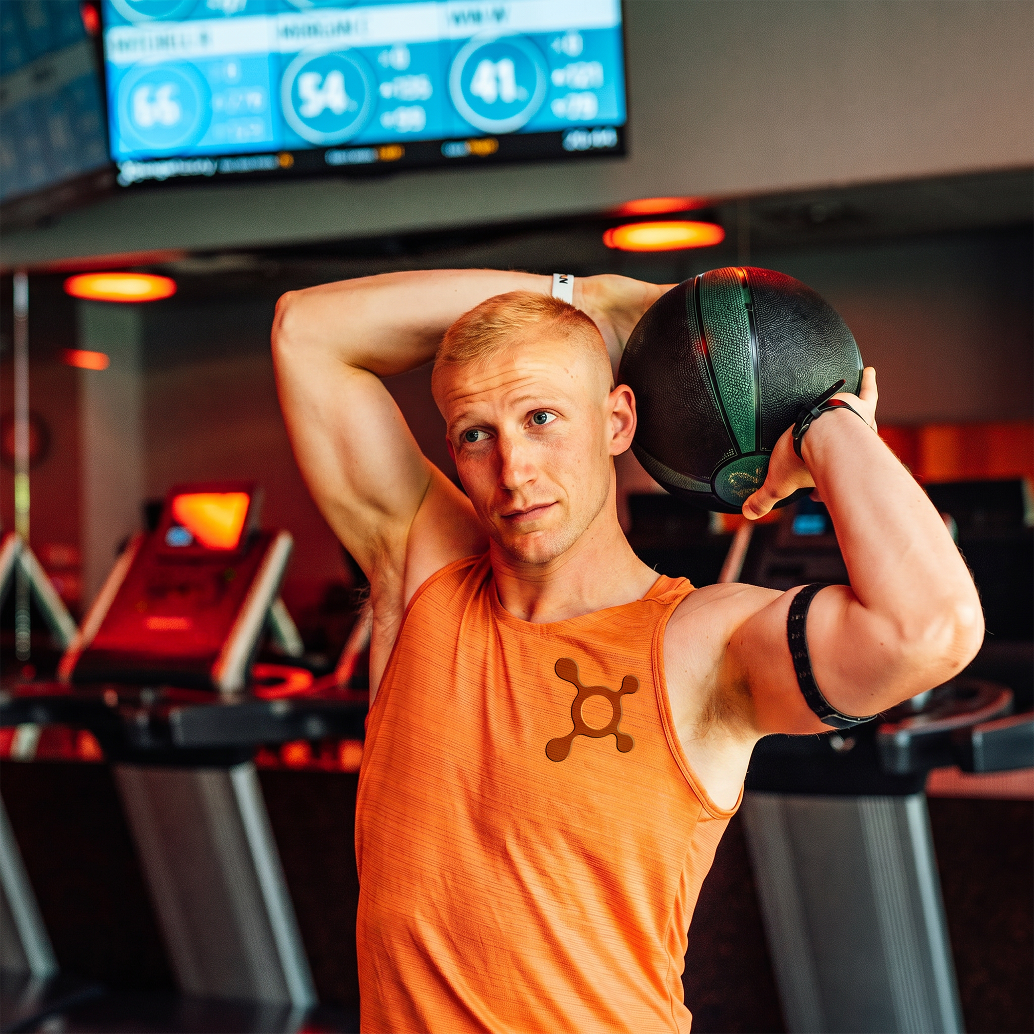 A young man with short blonde hair and blue eyes is lifting a black medicine ball overhead, in a gym with bright lighting and red accents. The man is wearing an orange tank top and sports armbands.