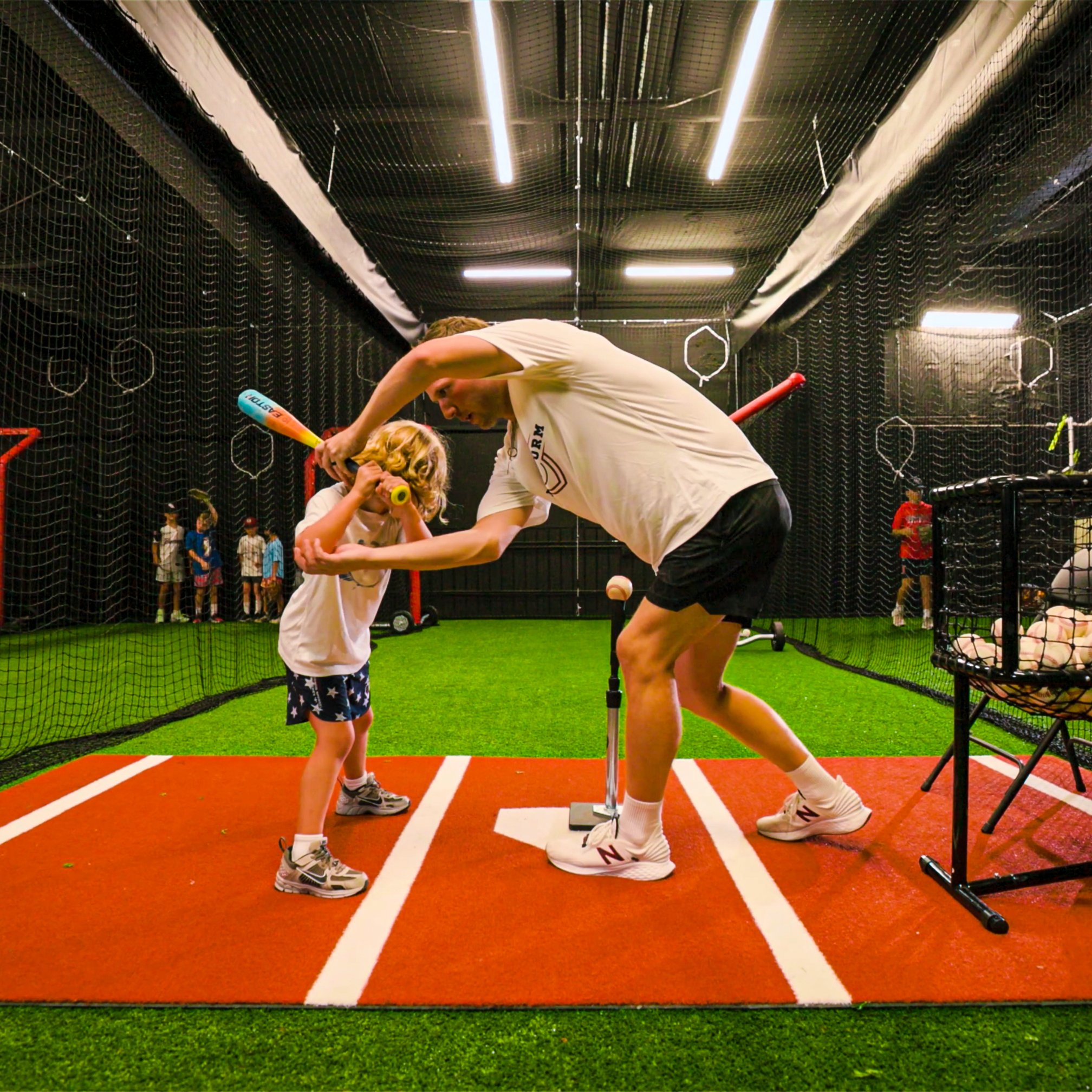 A baseball coach bending down to speak with a young boy holding a bat in an indoor batting cage with black netting, fluorescent lights, and sports equipment.