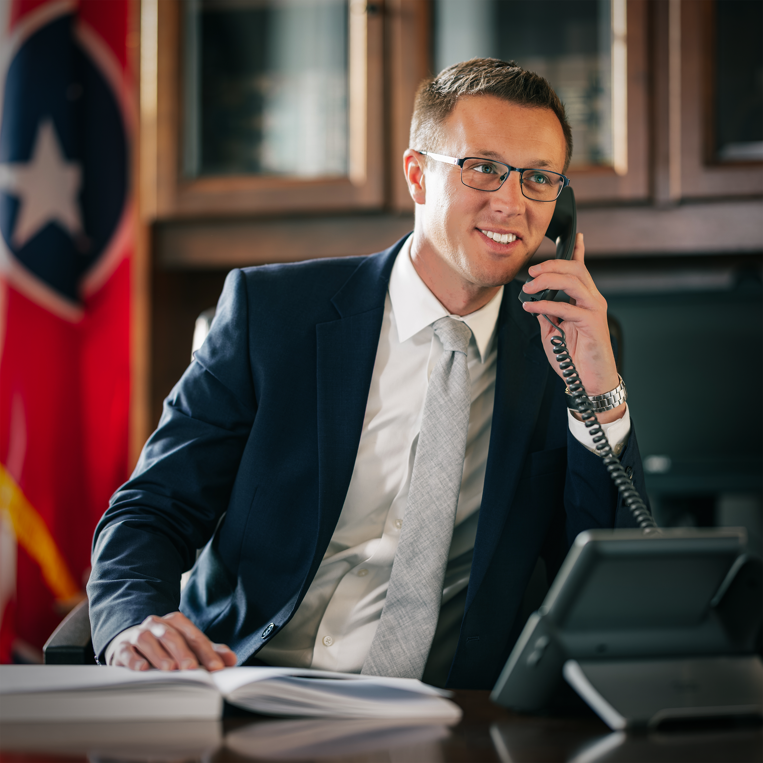 A young man with glasses in a business suit is sitting on a desk, smiling, and talking on a telephone. There is a U.S. flag in the background, indicating an office or governmental setting.