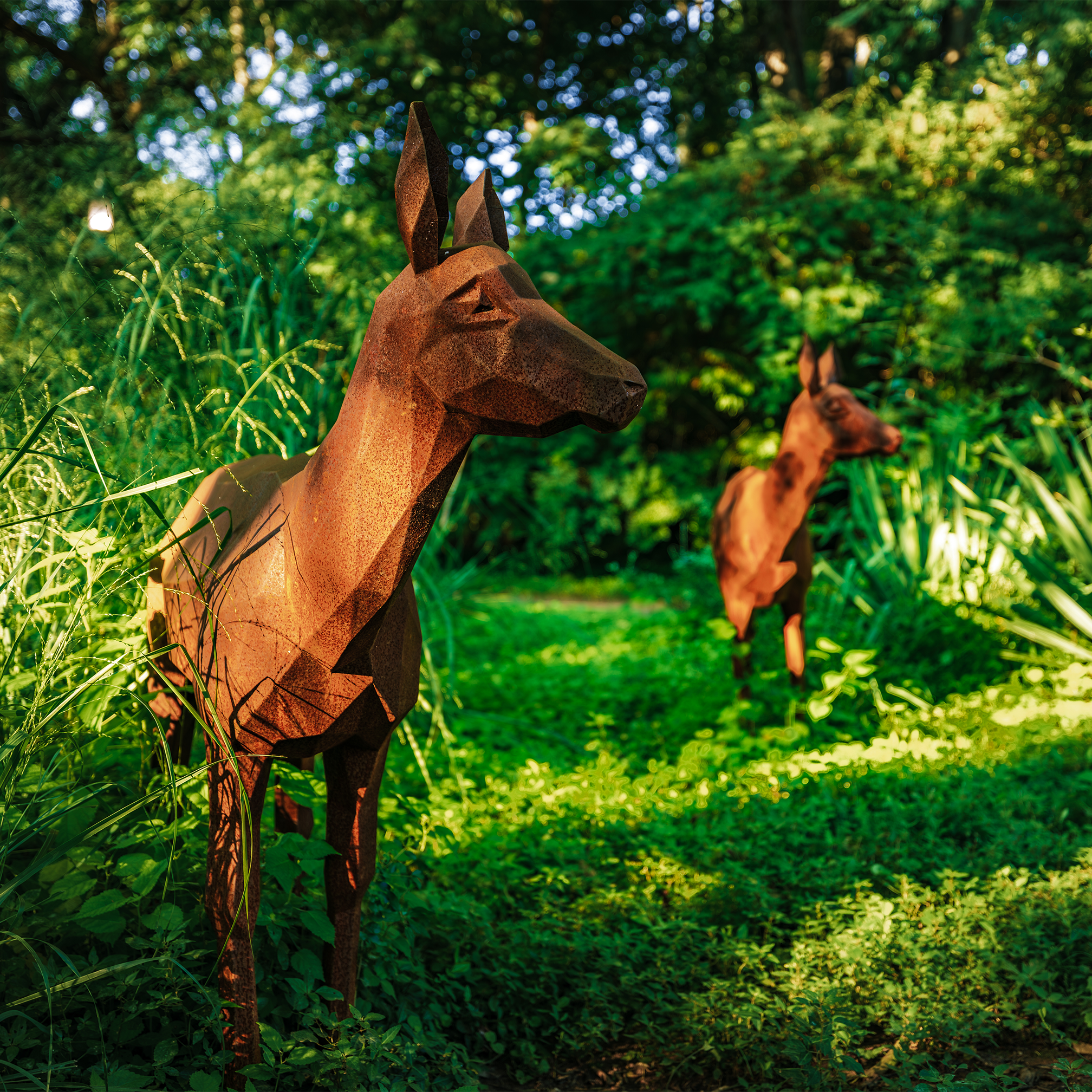 Rust-colored, geometrically stylized deer sculpture with pointed ears, set outdoors among green foliage, with trees and sunlight in the background.