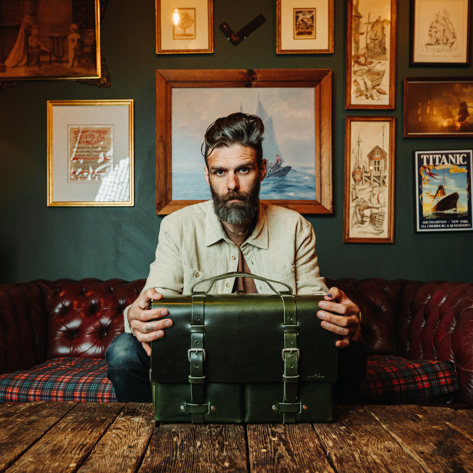 A man with a beard sitting on a red leather sofa, holding a vintage green suitcase in a room decorated with multiple framed pictures and artwork on the wall behind him. The room has a wooden floor and a dark green wall.
