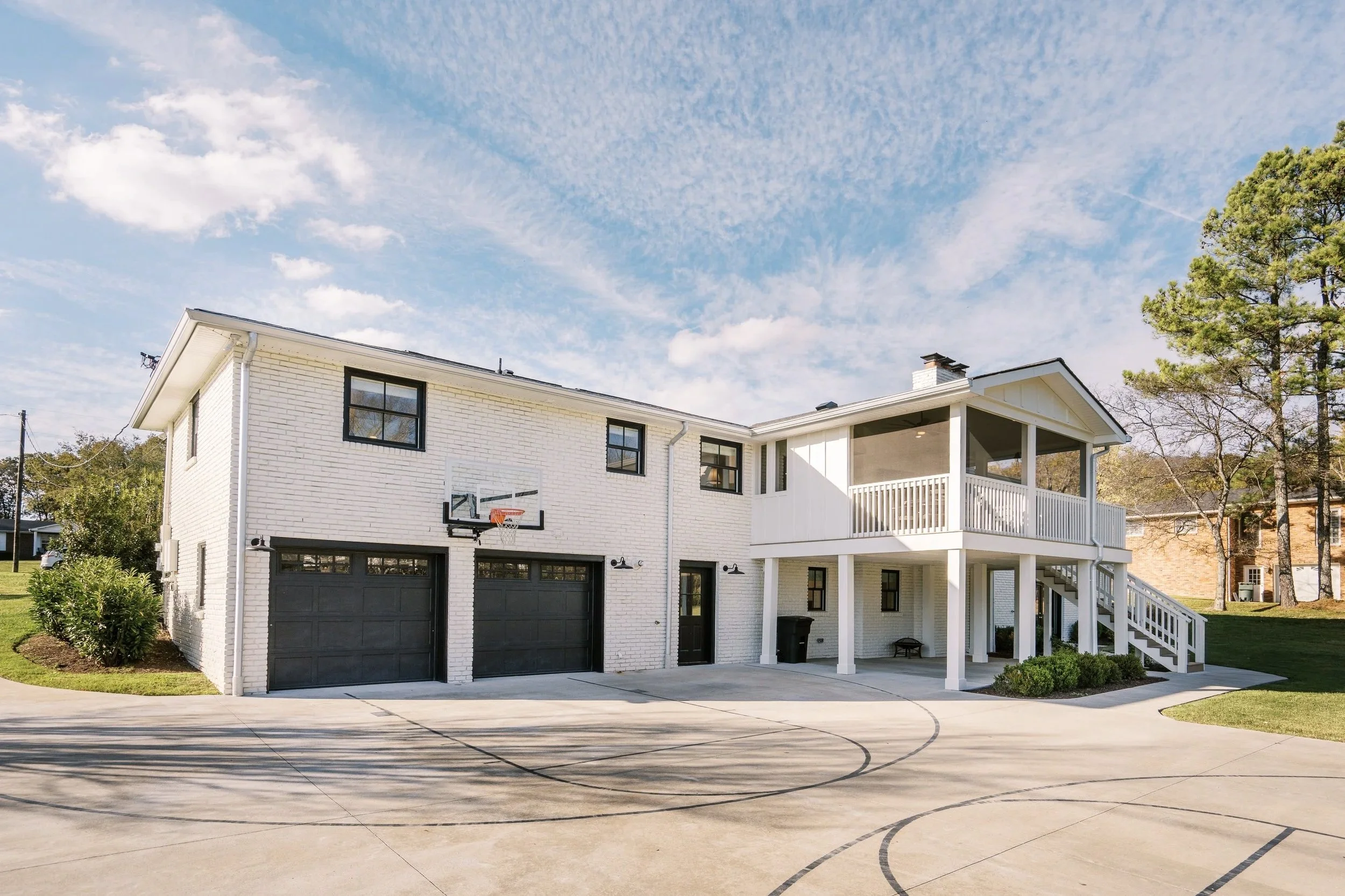 A white two-story house with a basketball hoop on the garage, a driveway, a deck with stairs, and trees on a sunny day with partly cloudy sky.