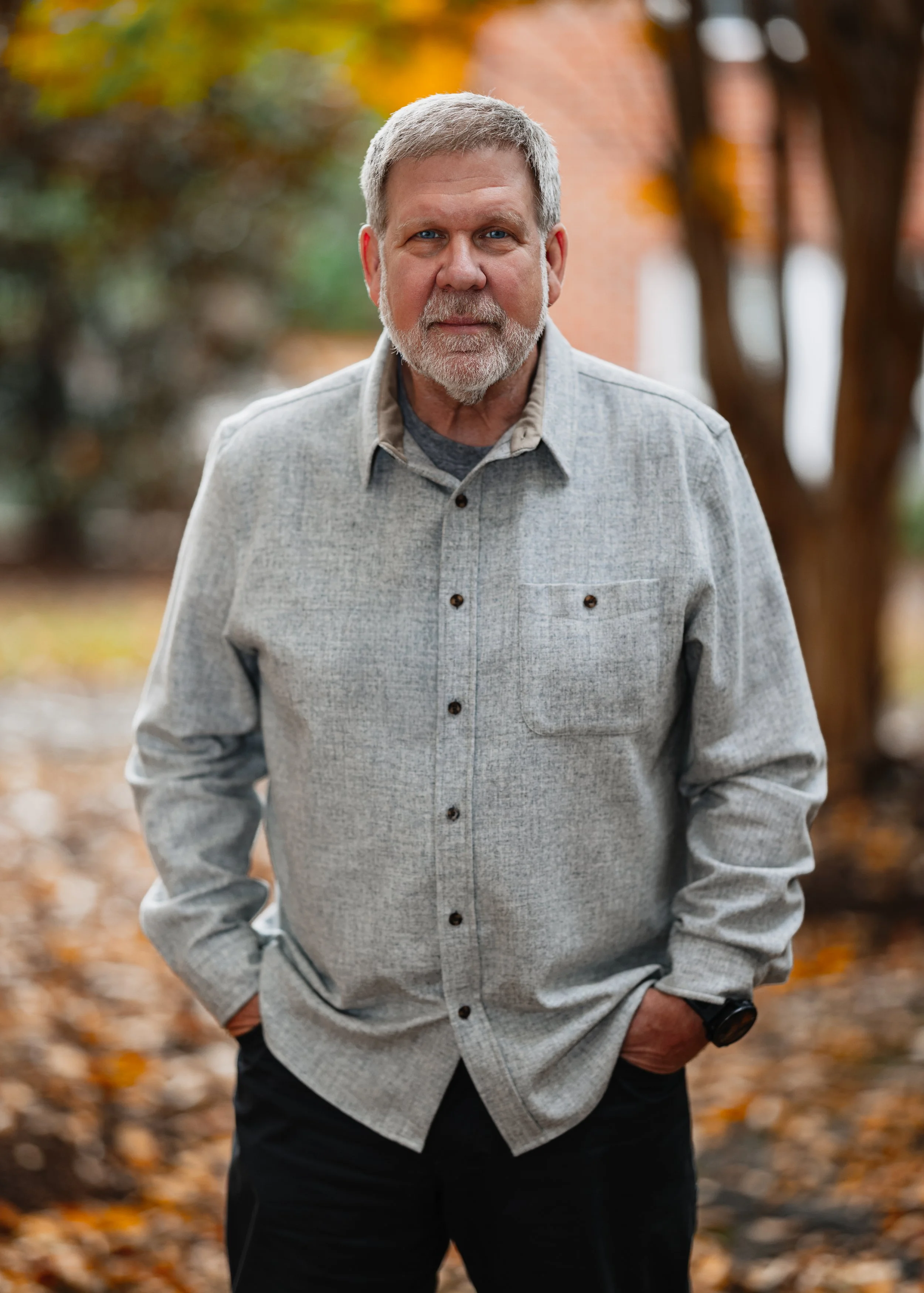 Portrait of Mark Holliday, founder of Pivot Construction, with gray hair and a beard wearing a light gray button-up shirt stands outdoors in a park with autumn leaves, with his hands in his pockets and a serious expression.
