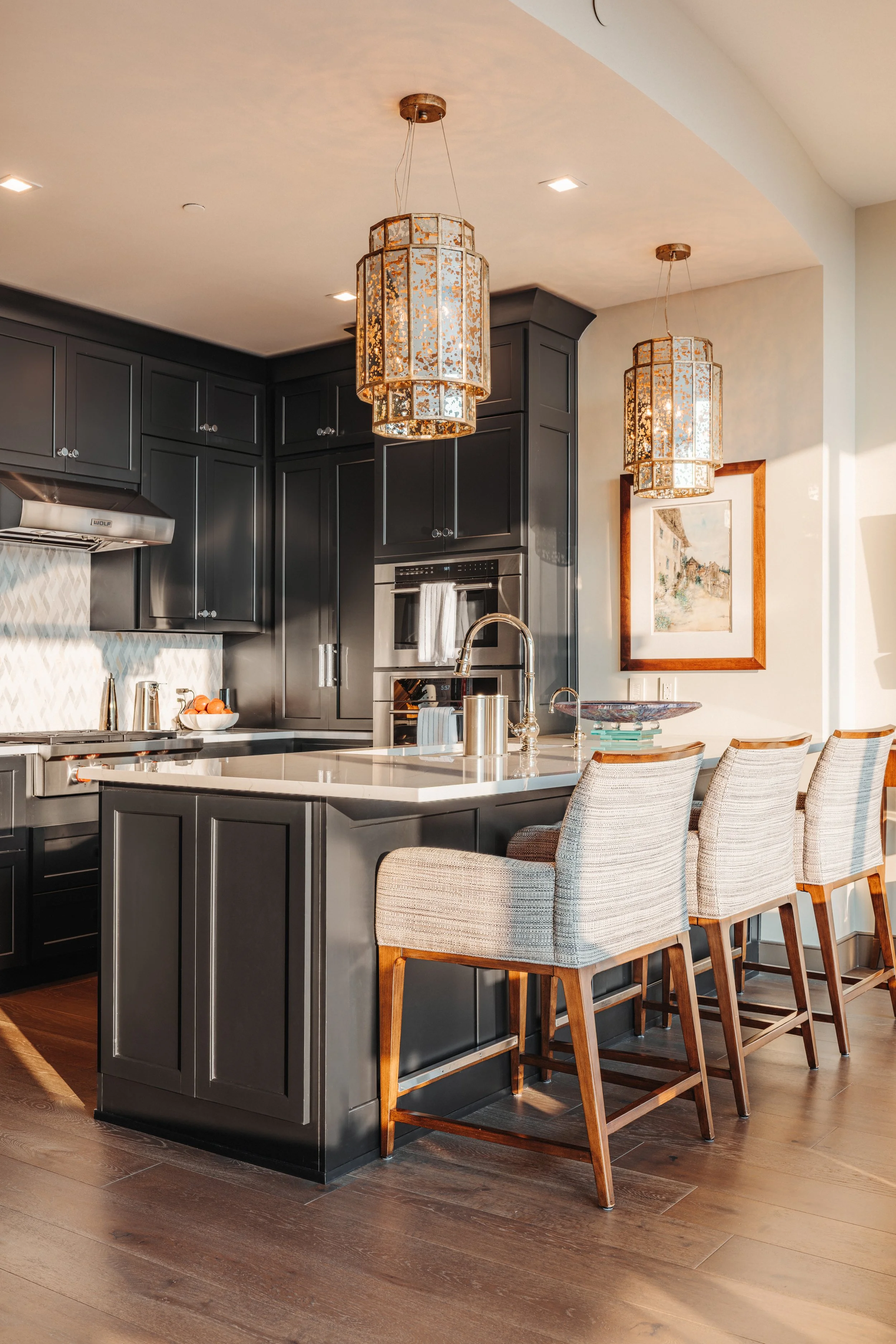A modern kitchen with a dark grey island, beige bar stools, black cabinetry, and gold pendant lights hanging from the ceiling. There's a painting on the wall near the window and wood flooring.