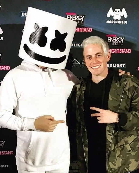 Two men stand side by side posing for a photo at a promotional event with a branded backdrop. The man on the left wears a large white marshmallow-shaped mask with black eyes and a wide smile, a white hoodie, and white pants, pointing to the right. Th