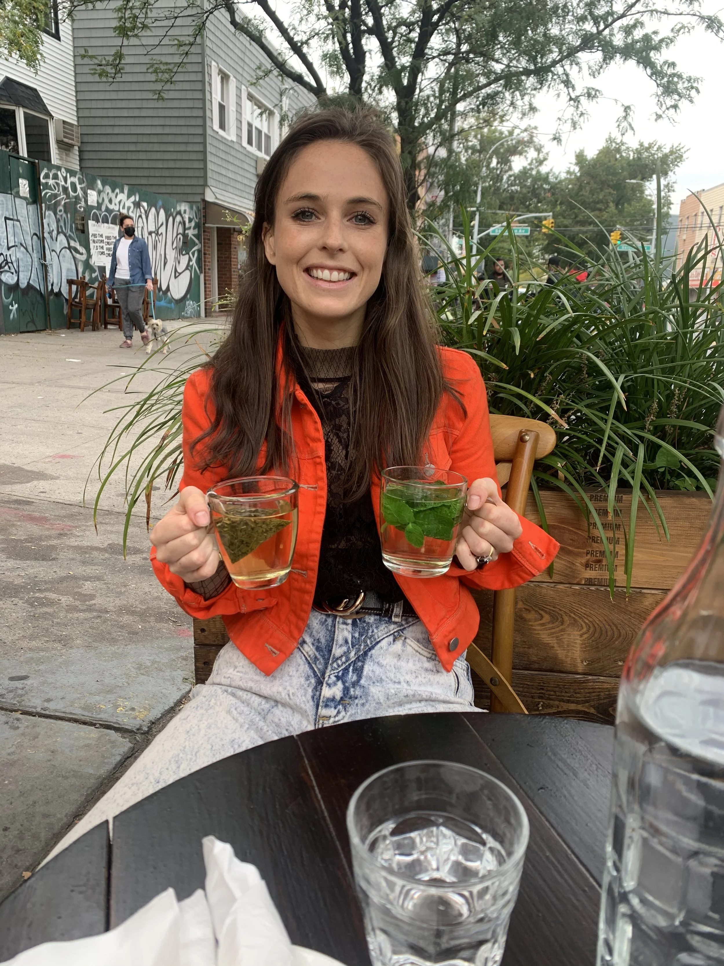 A young woman with long brown hair, wearing a red jacket and light-colored jeans, is sitting outdoors at a table, smiling, and holding two clear glasses of herbal tea with mint leaves. There is a large plant behind her, and other people are visible i