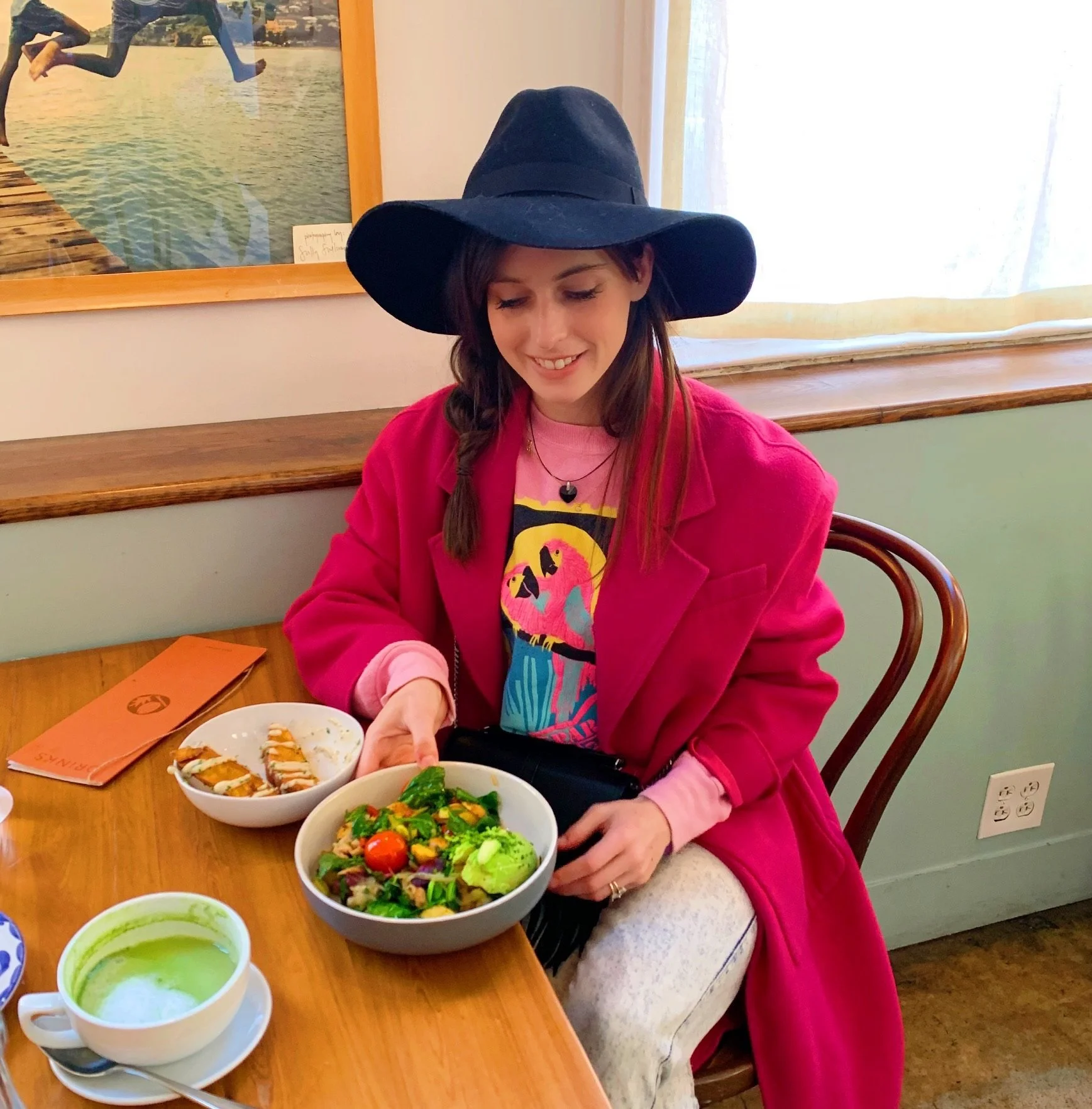 A woman wearing a large black hat, pink coat, and colorful shirt, sitting at a wooden table with a bowl of salad, a bowl of breaded food, and a cup of green soup.