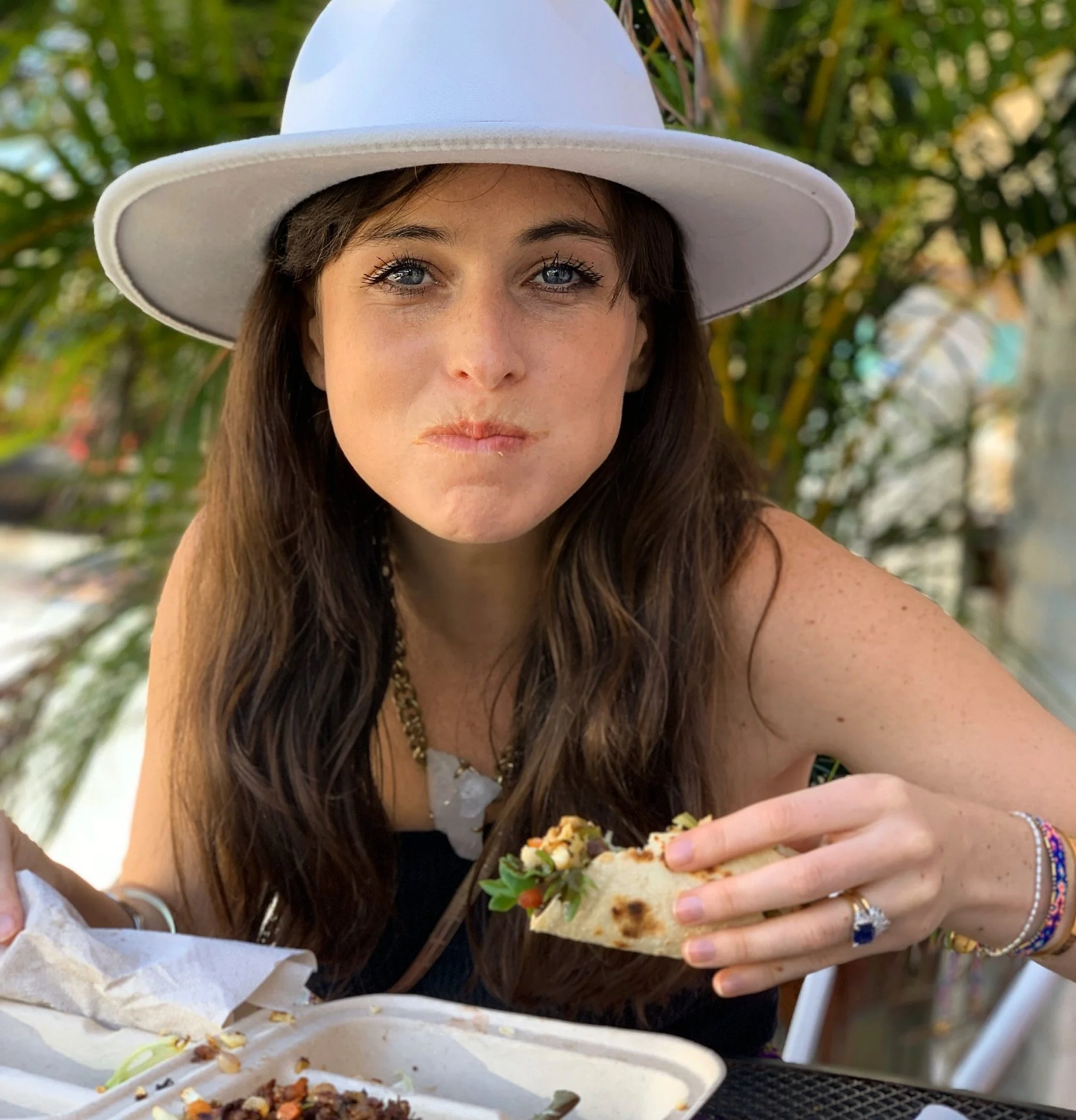 Young woman wearing a white hat eating a taco outdoors with palm trees in the background.