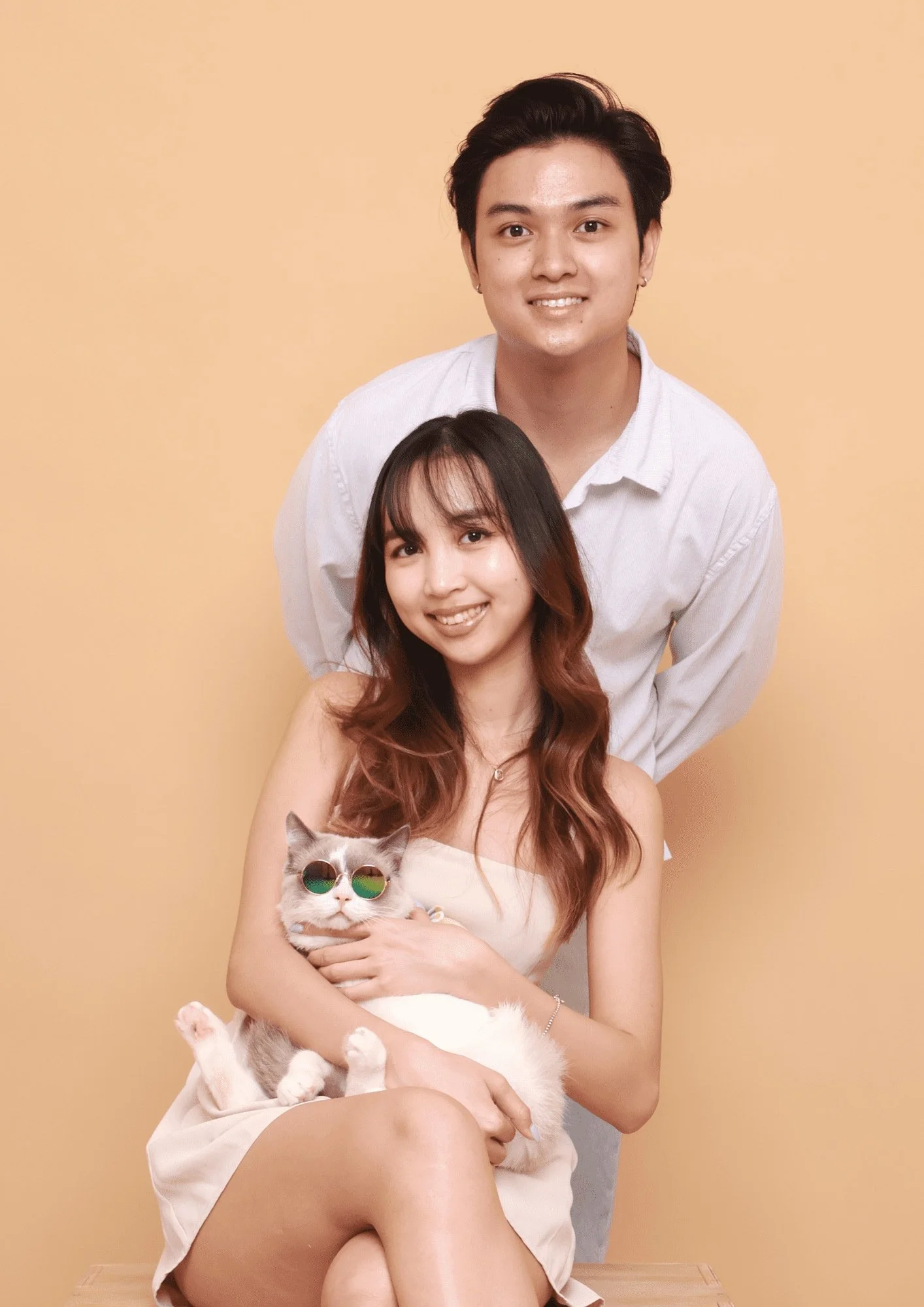 A smiling woman holding a cat with sunglasses, sitting in front of a man against a beige background.