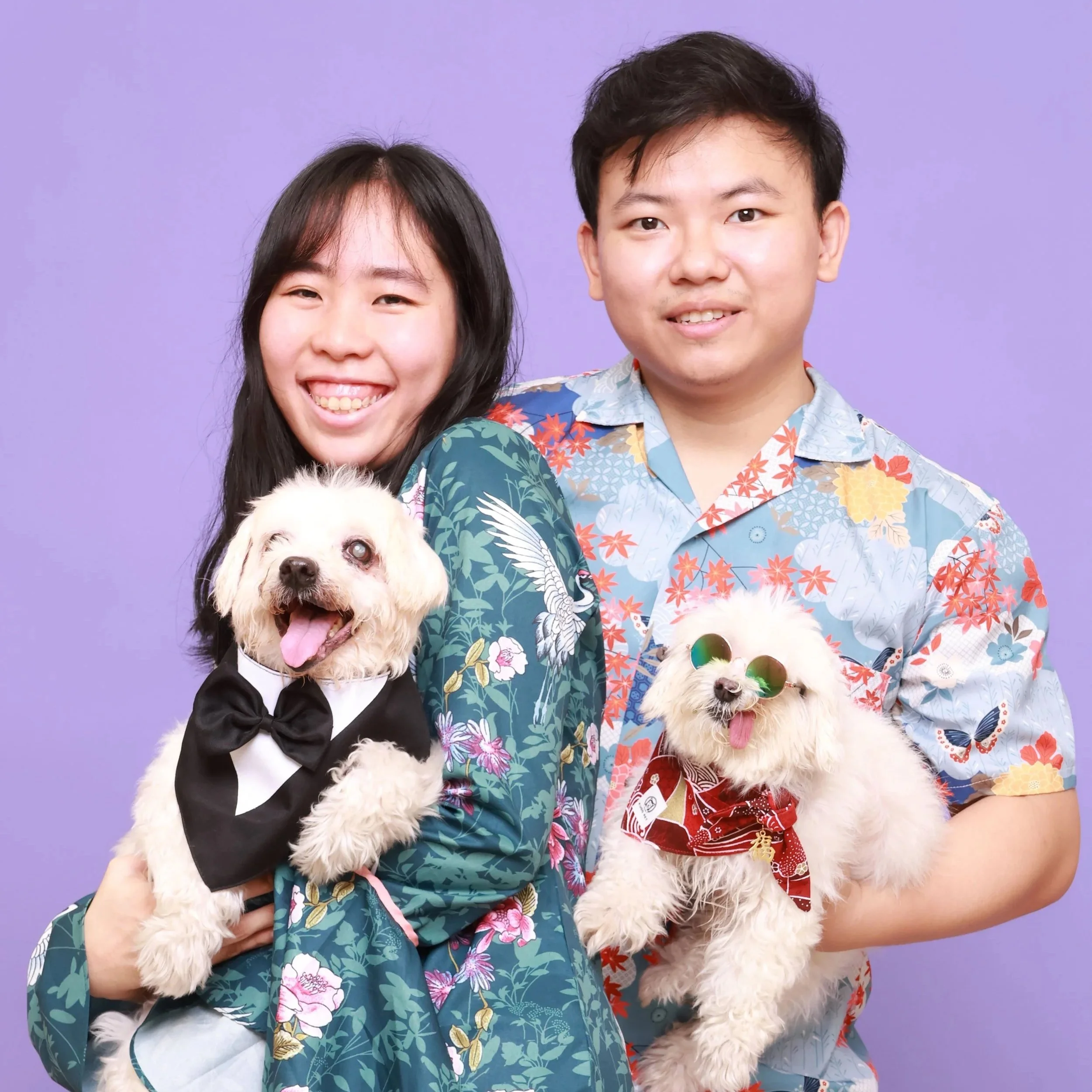 A smiling young Asian couple holding two small dogs in summer clothes against a purple background.