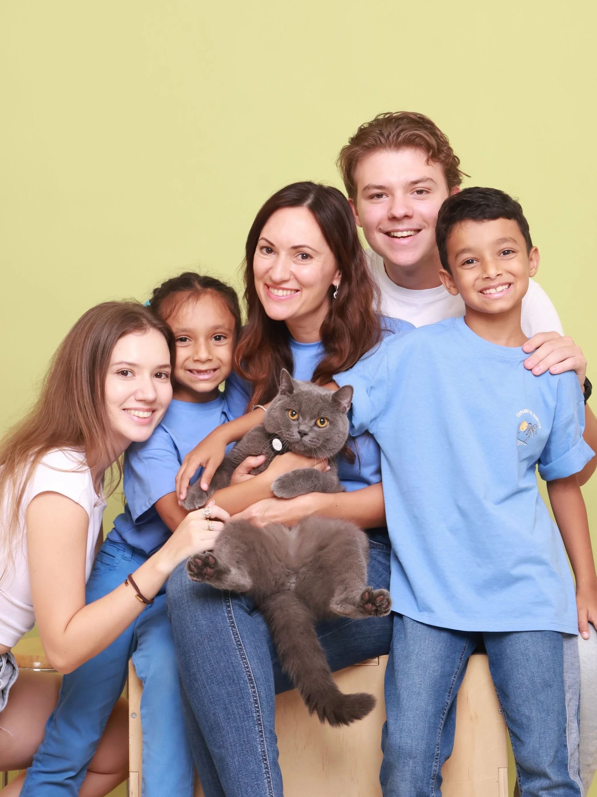 Group of five children and a woman holding a gray cat, smiling against a light green background.