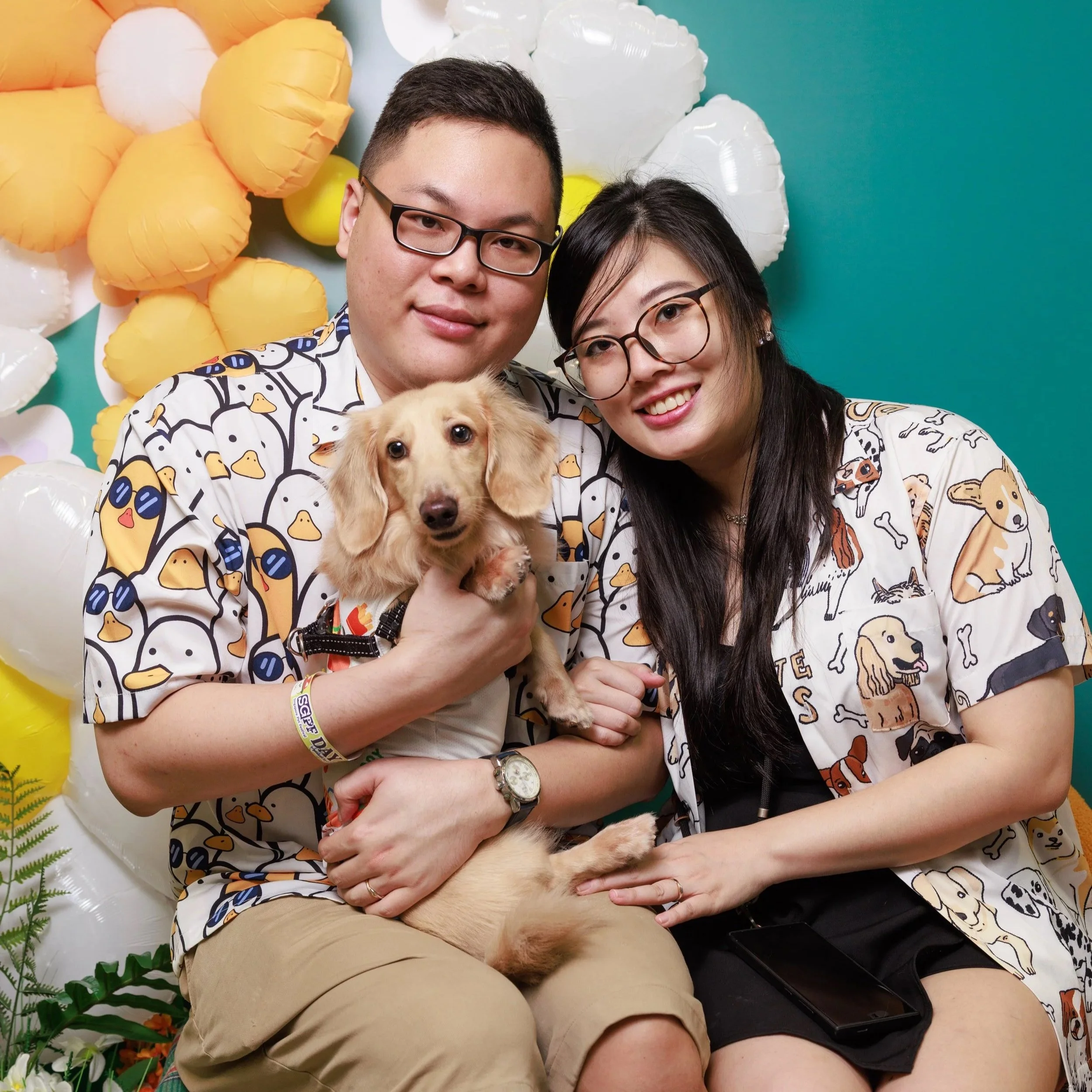 A happy couple dressed in matching dog-themed shirts sitting together with their dog in front of a colorful balloon and flower backdrop.