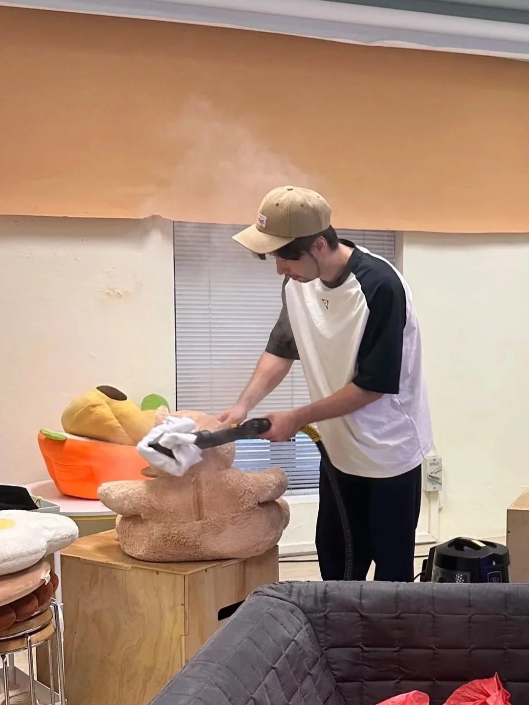 A person wearing a beige cap and a black and white t-shirt is vacuuming a plush teddy bear on a wooden table. The room has light-colored walls, a window with blinds, and various stuffed animals and objects around.
