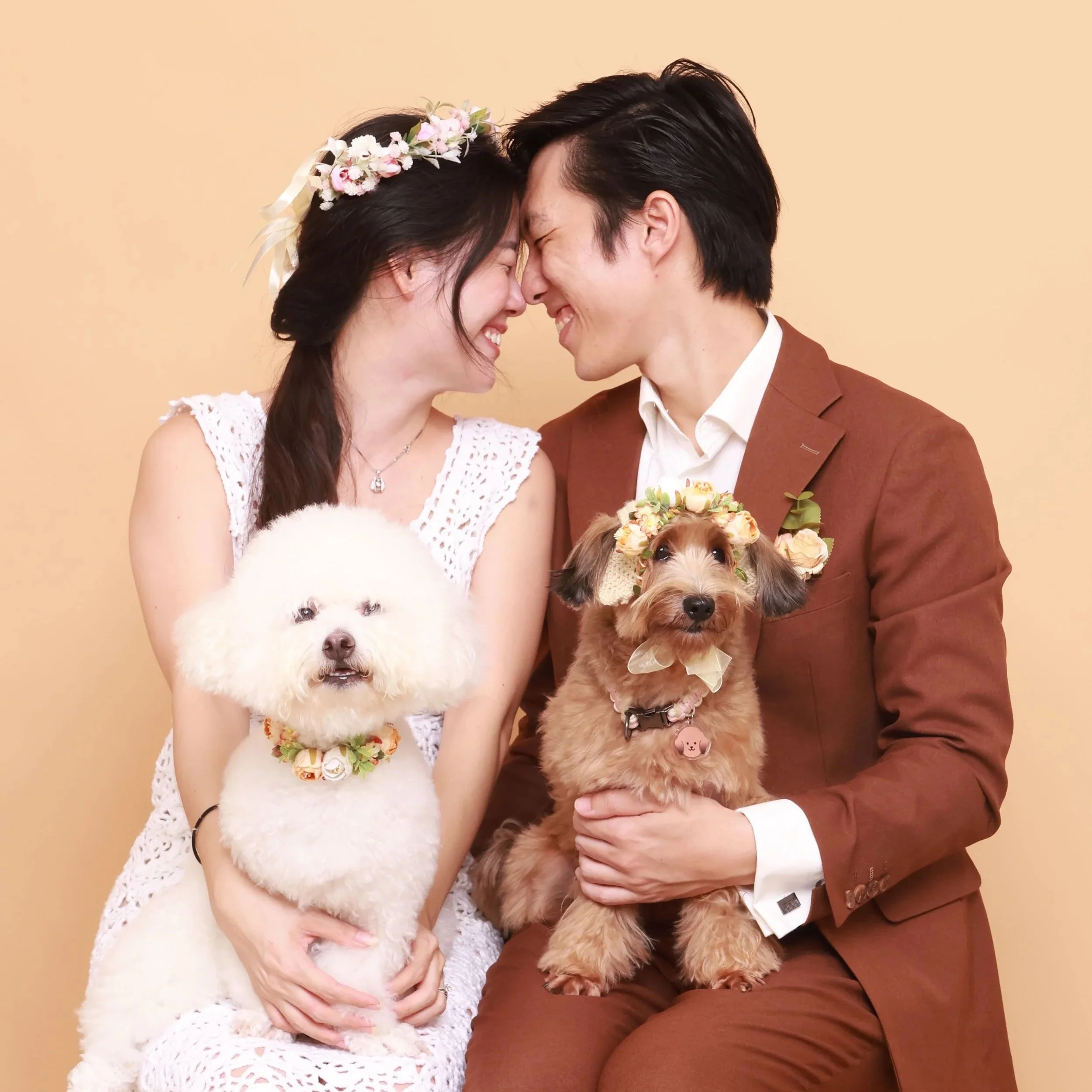 A smiling couple with two dogs, one white and one brown, wearing floral accessories, touching foreheads against a pastel background.