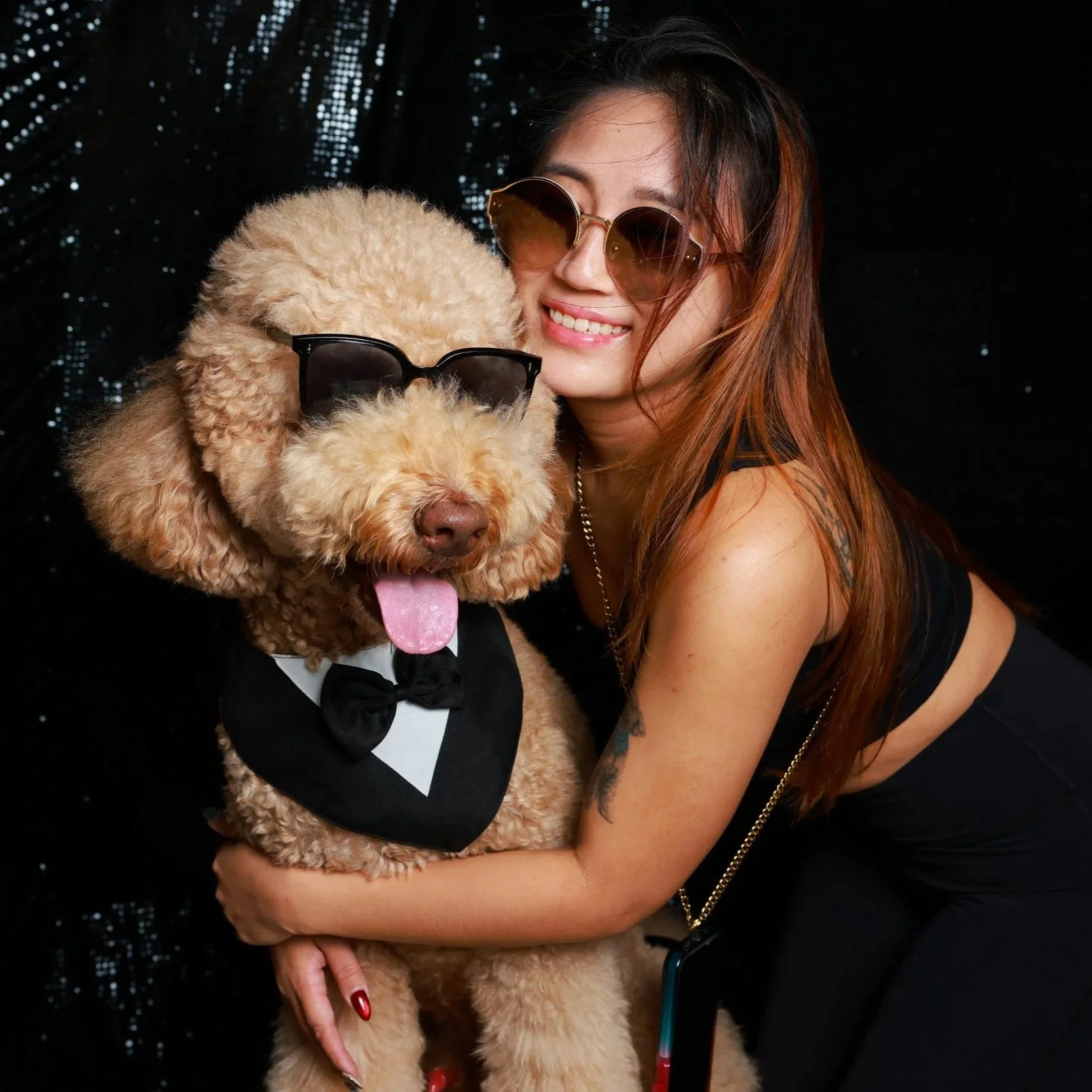 A woman in sunglasses and black clothing hugging a poodle wearing sunglasses, a tuxedo bib, and a bow tie, against a black background with sparkles.