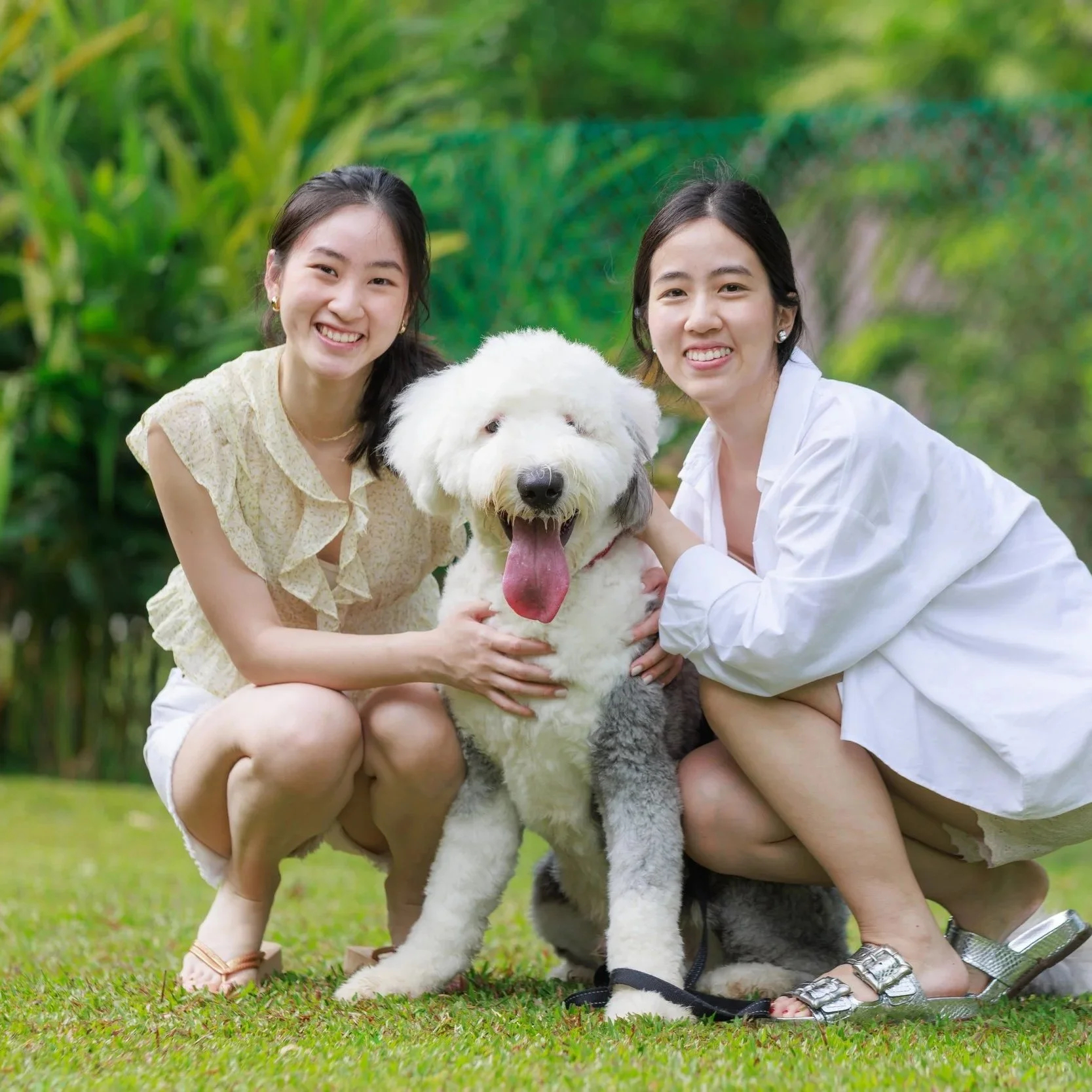 Two women smiling and hugging a fluffy white and gray dog with its tongue out, outdoors in a lush green garden.