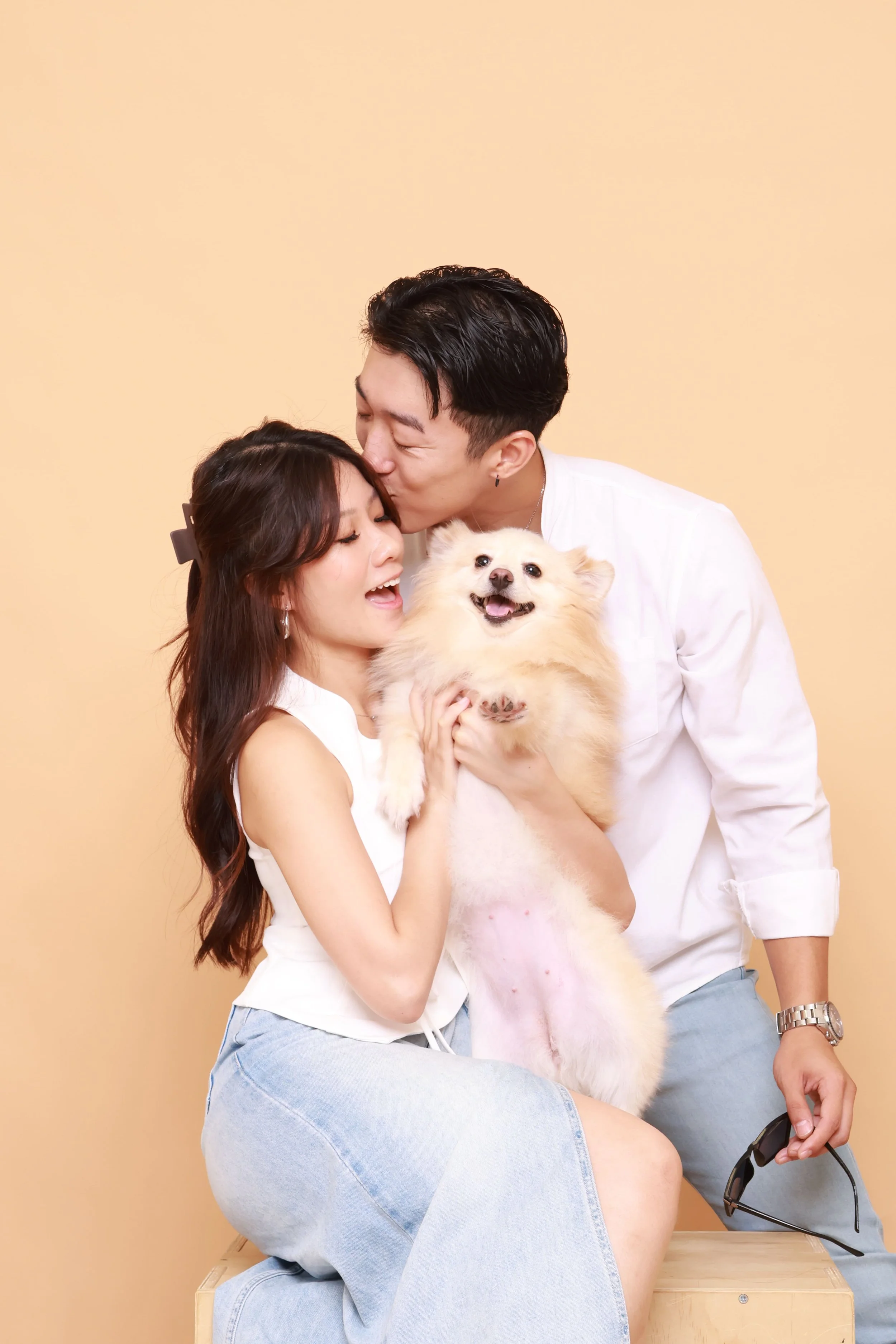 A couple with a fluffy, cream-colored dog against a plain beige background. The woman has long dark hair with a hair clip, and the man has short, styled black hair. The woman is sitting on a wooden box, holding the dog, and the man is standing behind her, kissing her forehead. The woman is wearing a white sleeveless top and light blue jeans, while the man is dressed in a white shirt and light-colored pants. The woman is smiling with her eyes closed, and the dog is smiling with its tongue out.