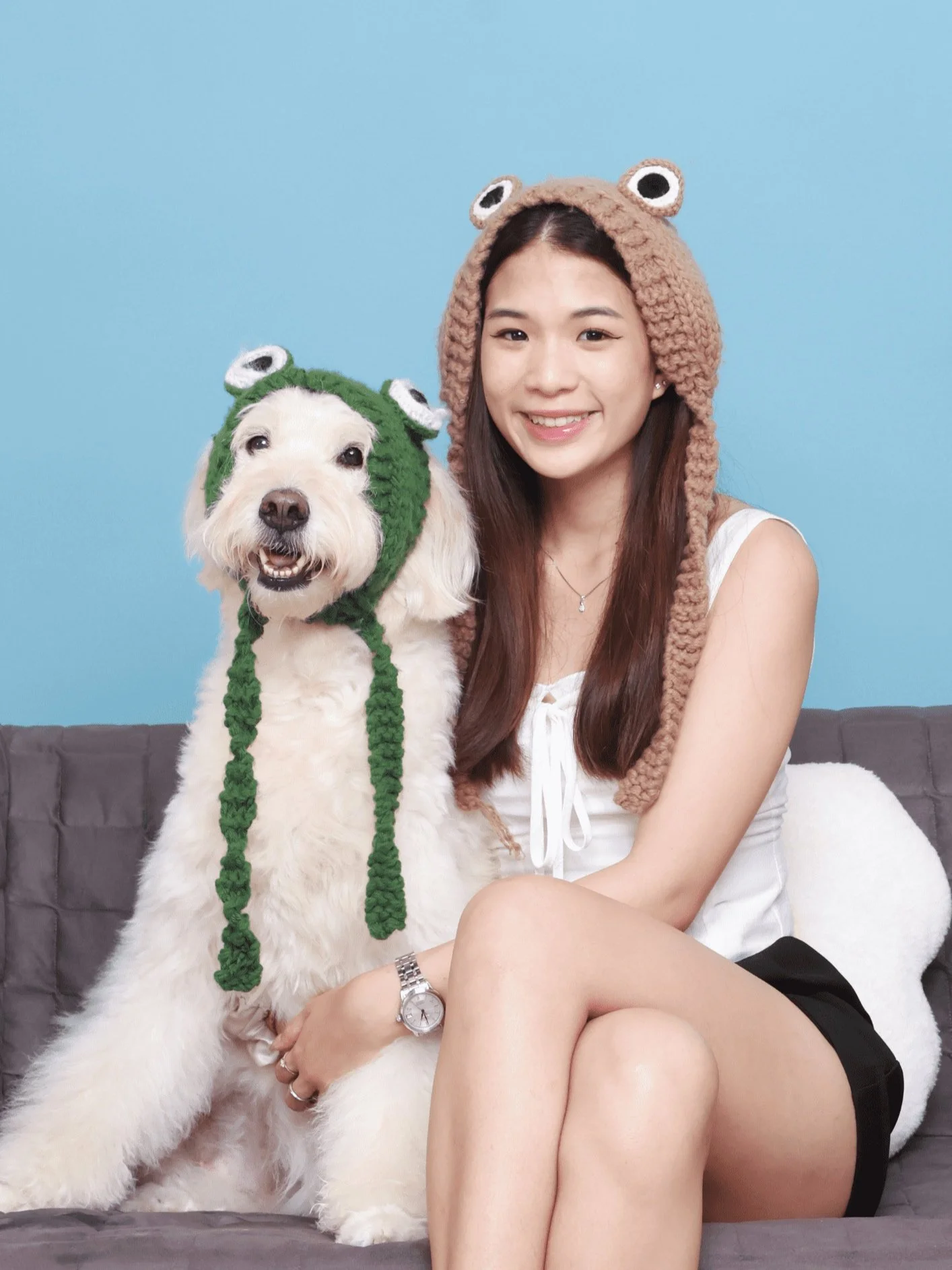 A woman sits on a sofa next to a fluffy white dog. Both are wearing knitted hats with animal ears; the woman has a brown hat, and the dog has a green hat. The background is light blue.