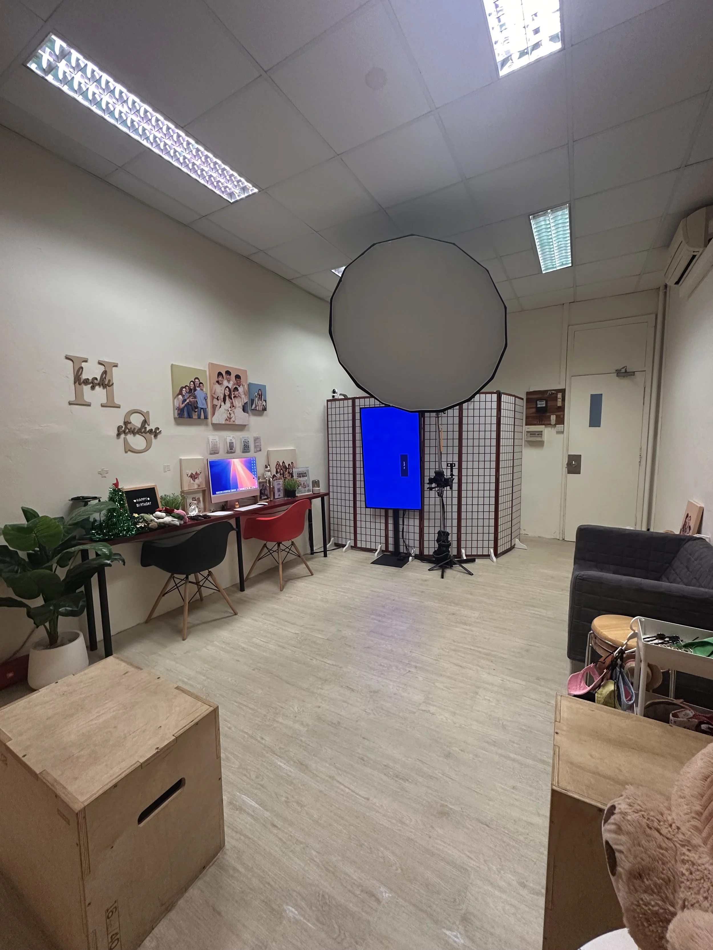 A photography studio setup with a large softbox, a photo backdrop screen, a digital monitor, a camera on a tripod, and various furniture and decorations suggesting a cozy work environment.