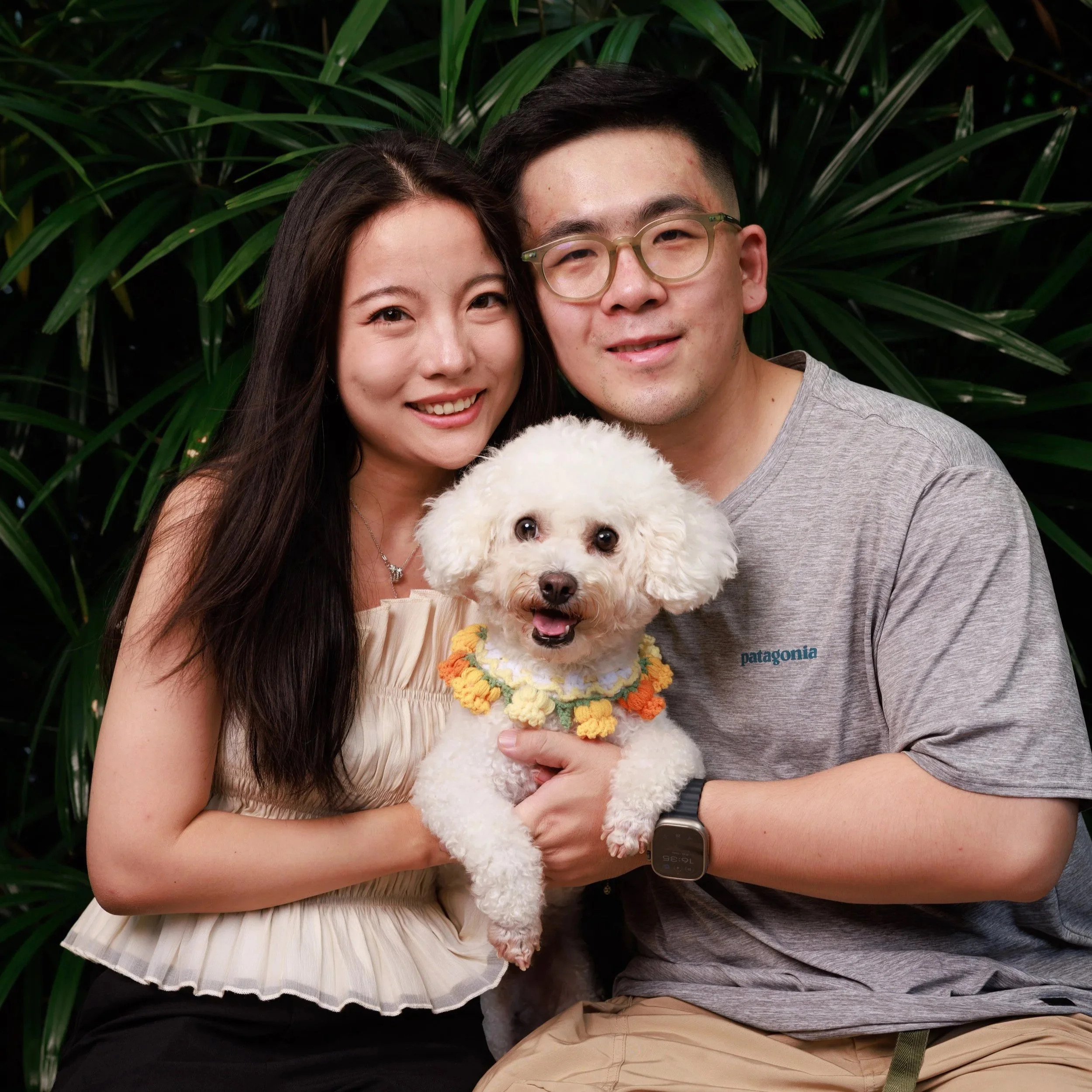 A smiling Asian woman and a smiling Asian man holding a small white poodle with a colorful flower collar, standing in front of green foliage.
