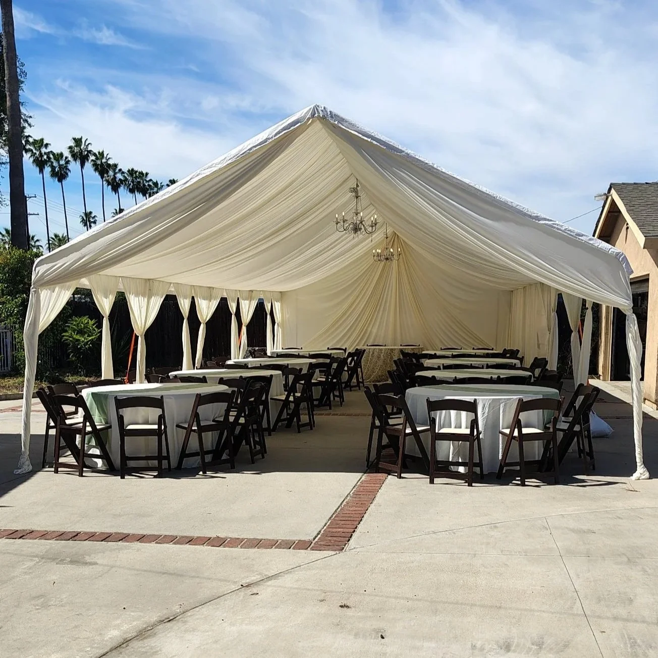 A large outdoor event tent with Ivori drapes and chandeliers, set up with round tables and brown chairs