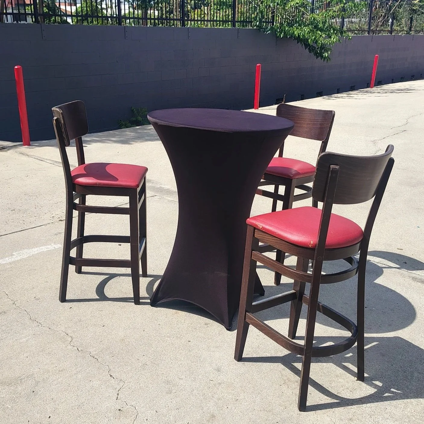 An outdoor patio scene with a tall round table covered with a black cloth, surrounded by four chairs with red cushions, on a concrete surface with cracks. In the background, there is a black wall with a metal fence on top and some green foliage.