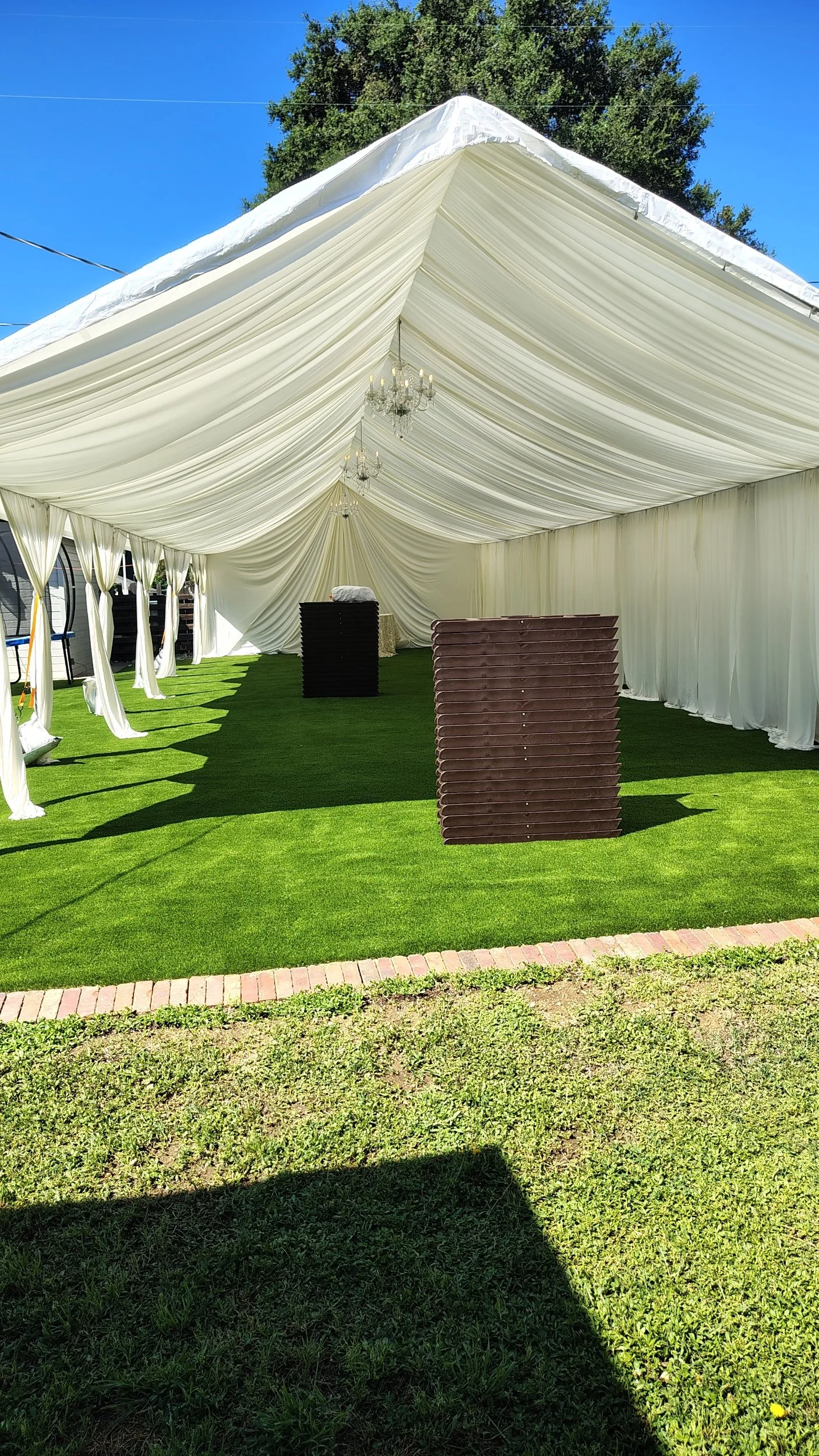 Outdoor event tent with white drapes and chandeliers, set up on a grassy area