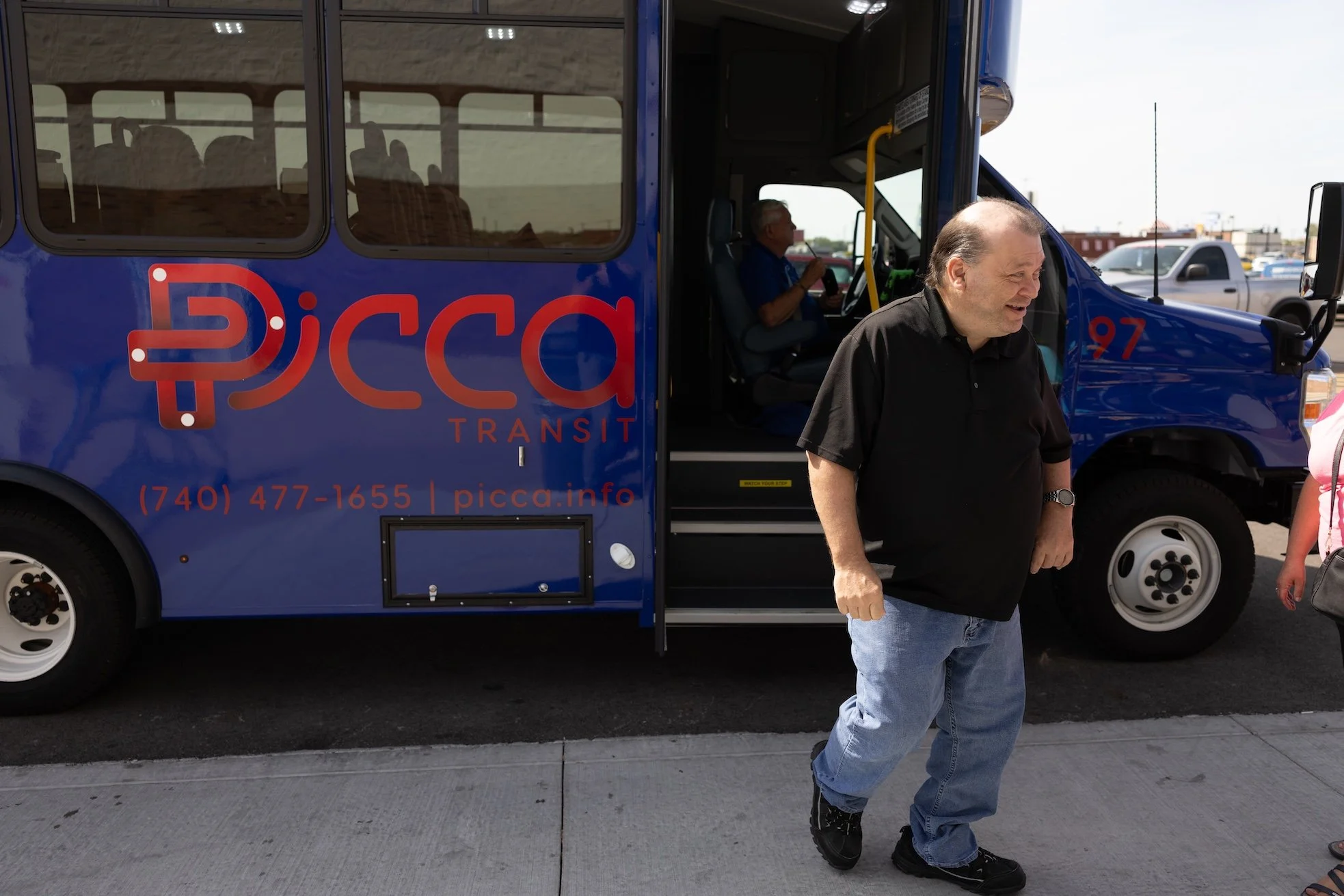 A man in a black shirt and blue jeans walking past a blue transit bus with the logo 'picca Transit' and a phone number. The bus has a driver inside and there are other vehicles in the background.