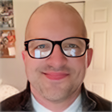 A man with glasses smiling indoors with a door and photos in the background.