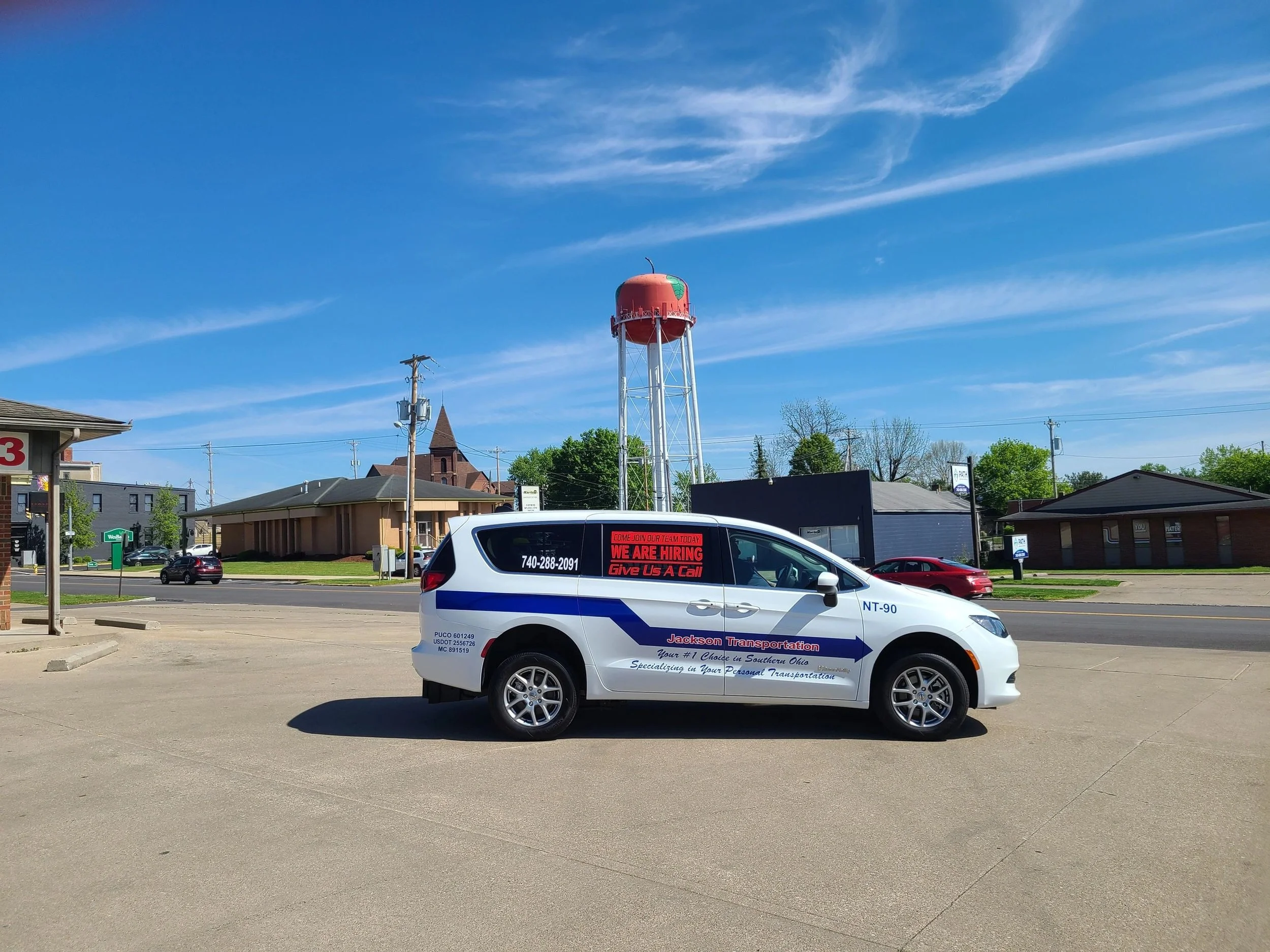 A Jackson Transportation company vehicle parked in an empty lot with a red water tower, buildings, trees, and a blue sky with wispy clouds in the background.