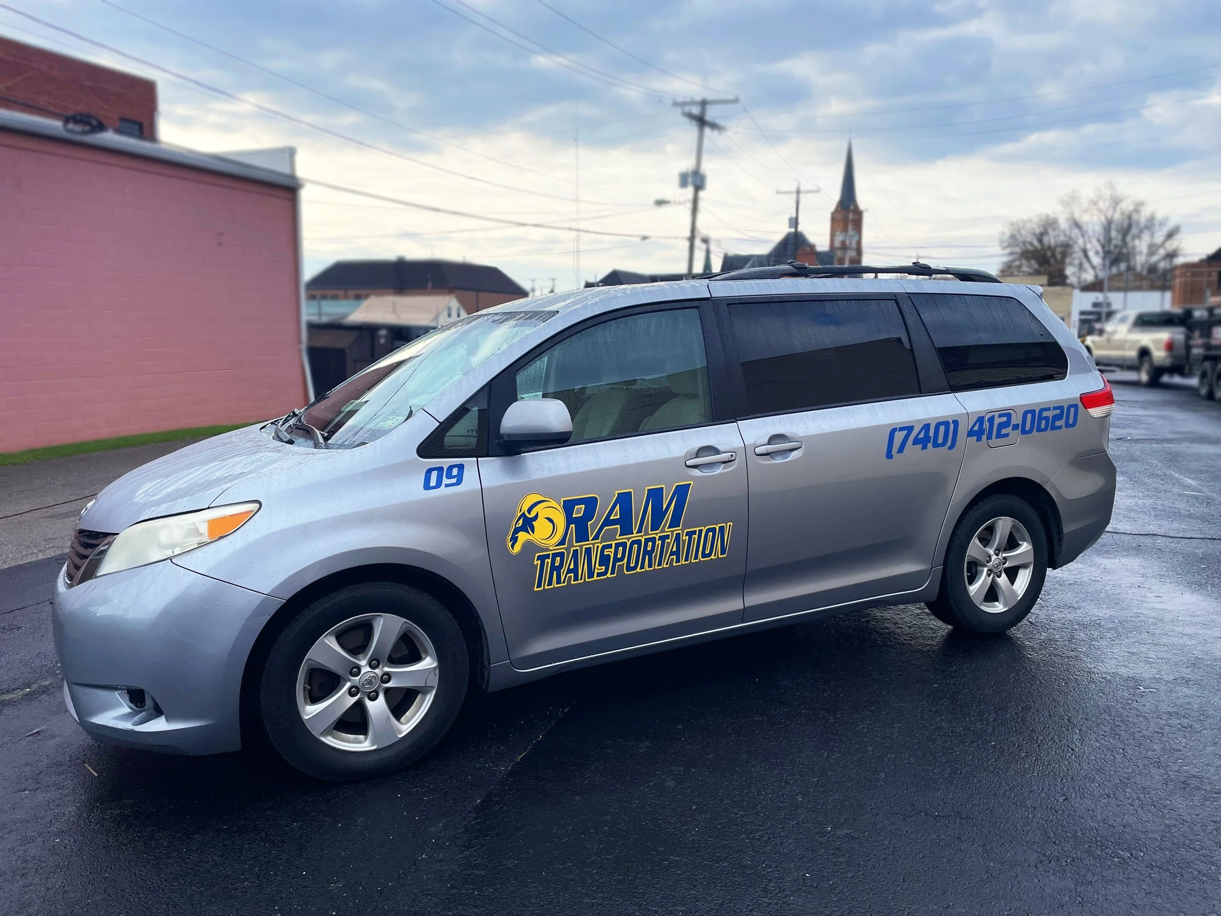 Silver minivan with RAM Transportation logo, blue and yellow, parked on wet asphalt in a lot with buildings and church steeple in background.