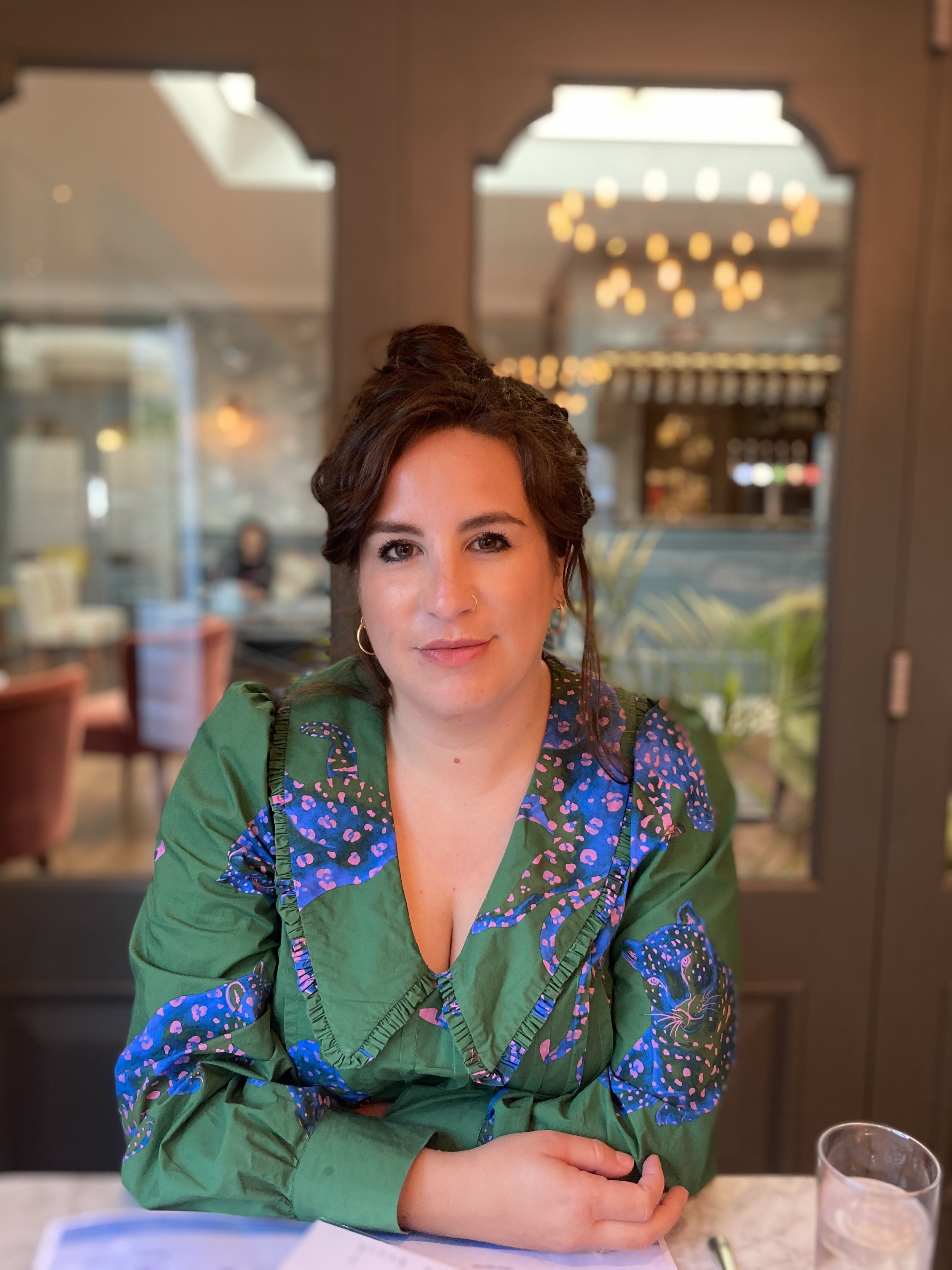 A woman with short dark hair and a nose ring, wearing a colorful patterned green jacket, sitting at a table in a restaurant or café, with blurred background including hanging lights.