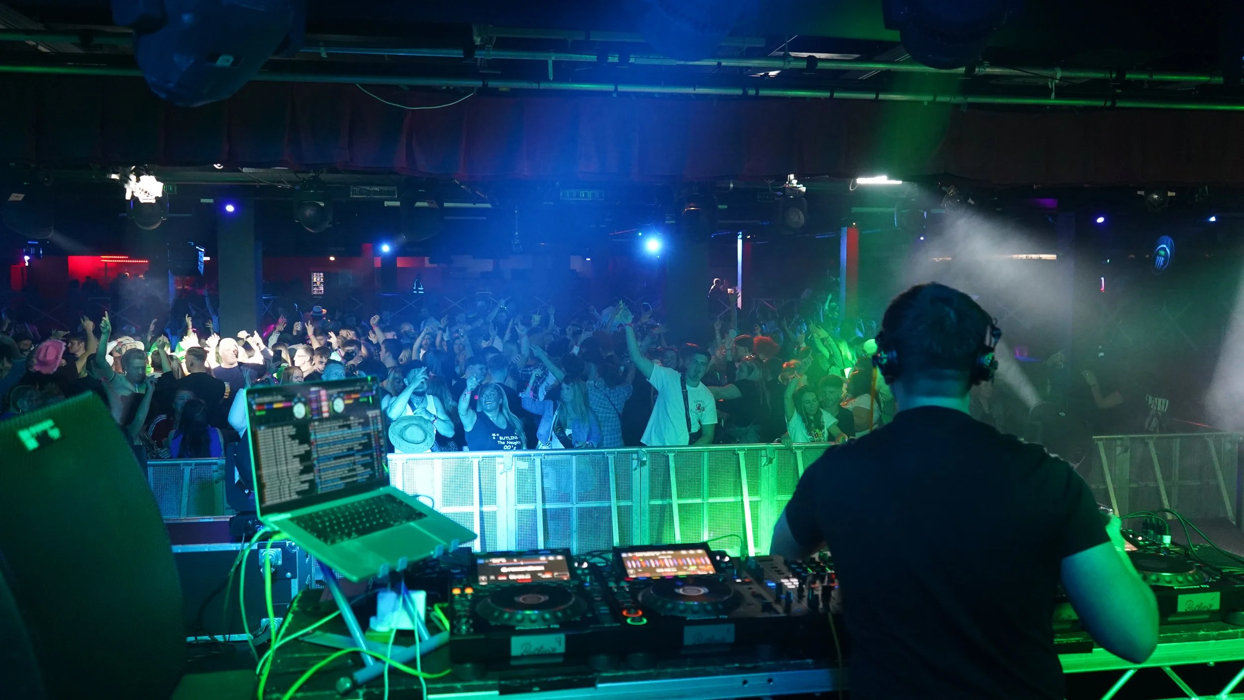 DJ performing at a club with a crowded dance floor, colorful lighting, and fog effects.