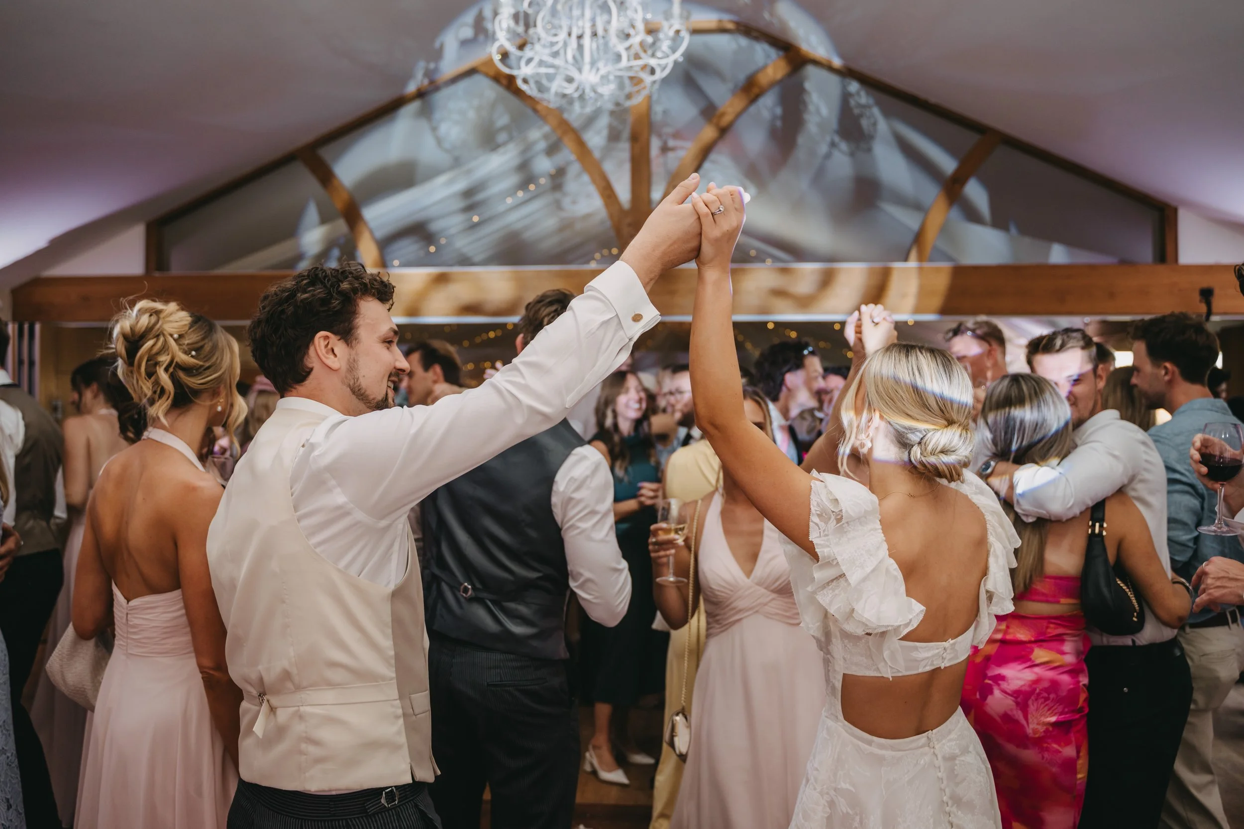 Couple dancing and holding hands at wedding reception with other guests dancing and celebrating in the background inside a decorated venue.