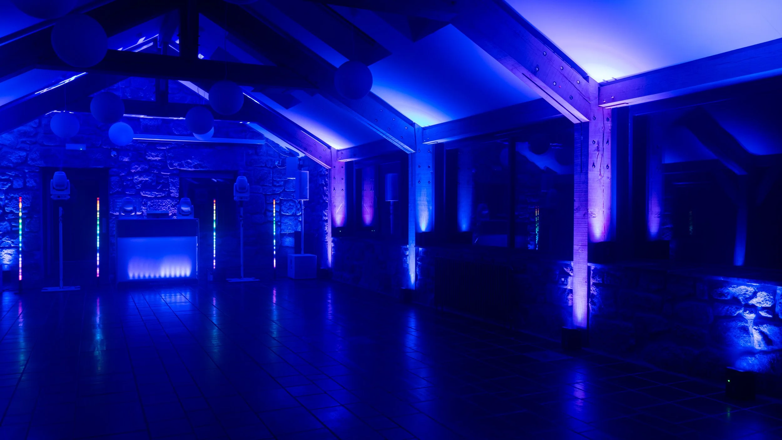 Interior of a restaurant or event space with colorful lighting. The room is decorated with red and green spotlights and has tables with people sitting, possibly for a gathering or event. A Christmas tree is visible, suggesting a festive atmosphere.