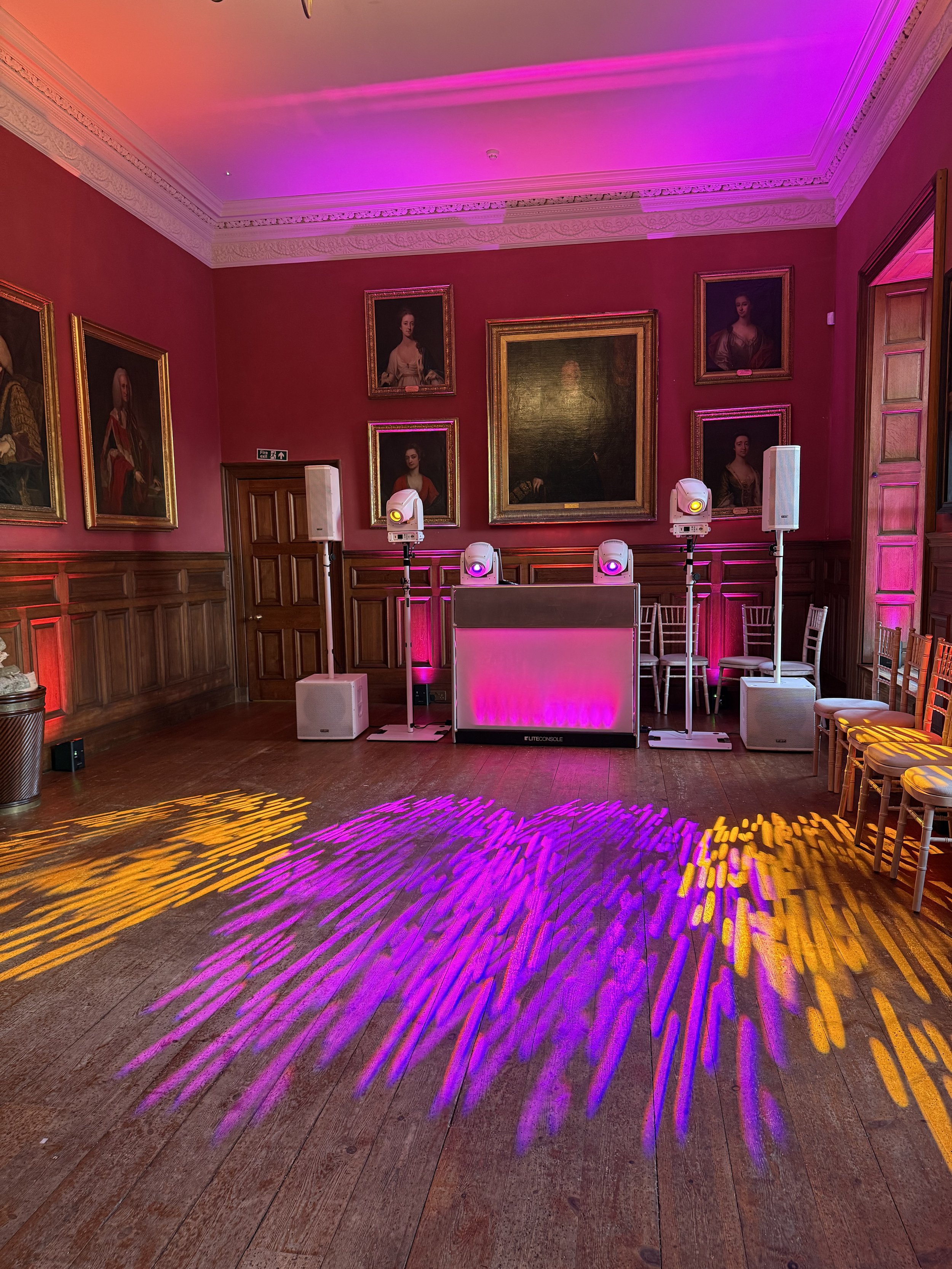 Elegant room with red walls, a fireplace, chandeliers, ornate ceiling patterns, and framed portraits on the walls.