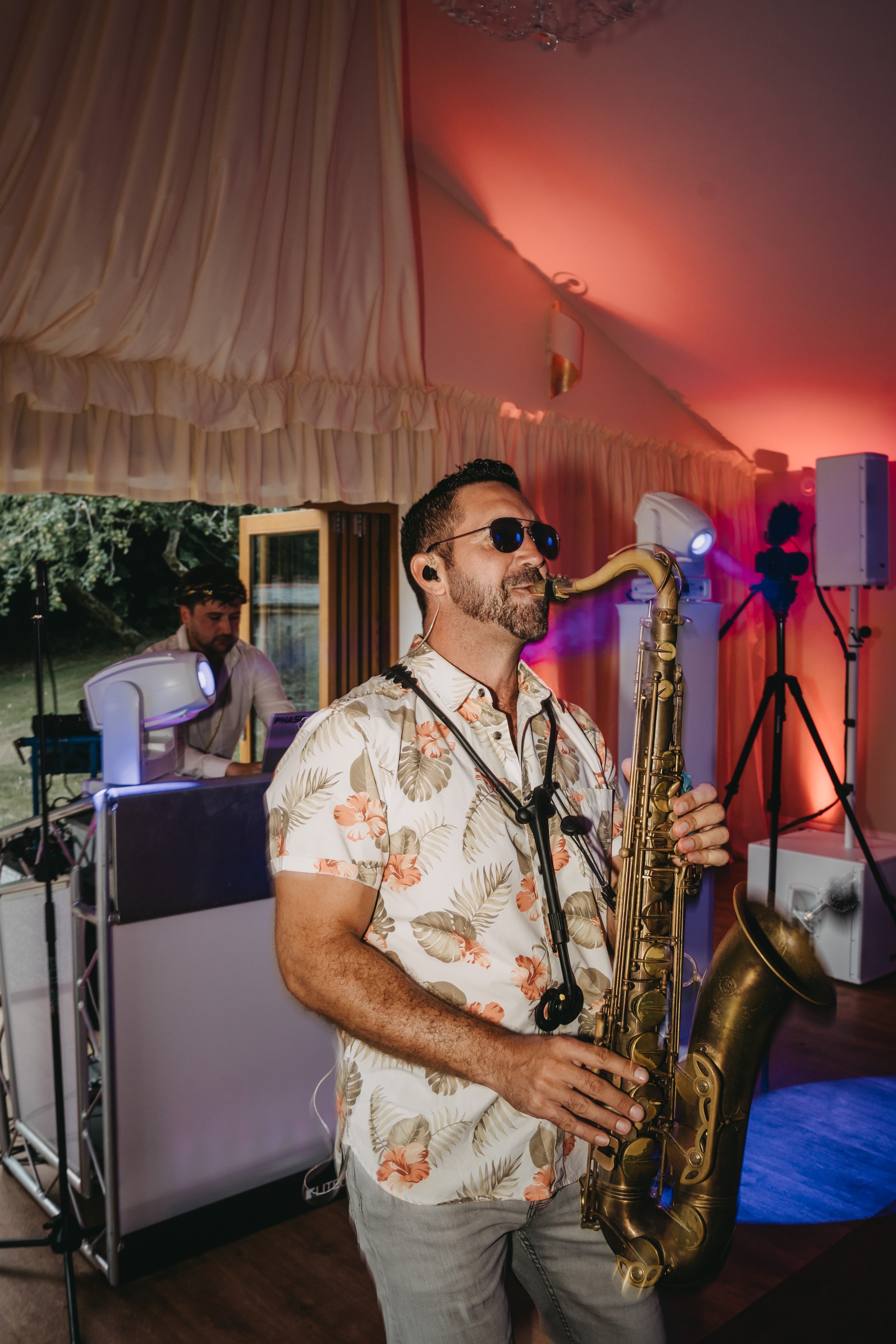 A man playing a saxophone at a party or event with a DJ in the background and colorful lighting.