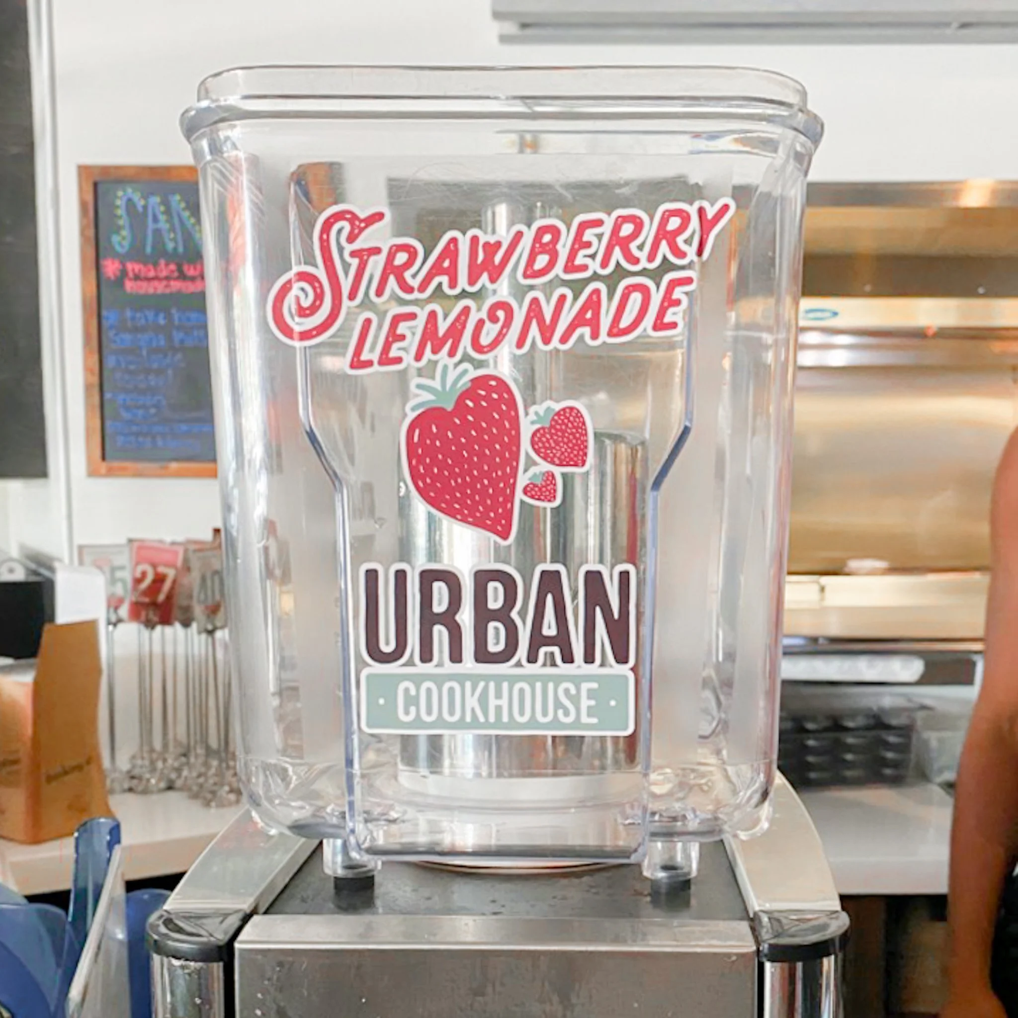 A clear plastic dispenser with a label that reads 'Strawberry Lemonade' and a logo for Urban Cookhouse, featuring strawberries and playful text, likely containing strawberry lemonade.
