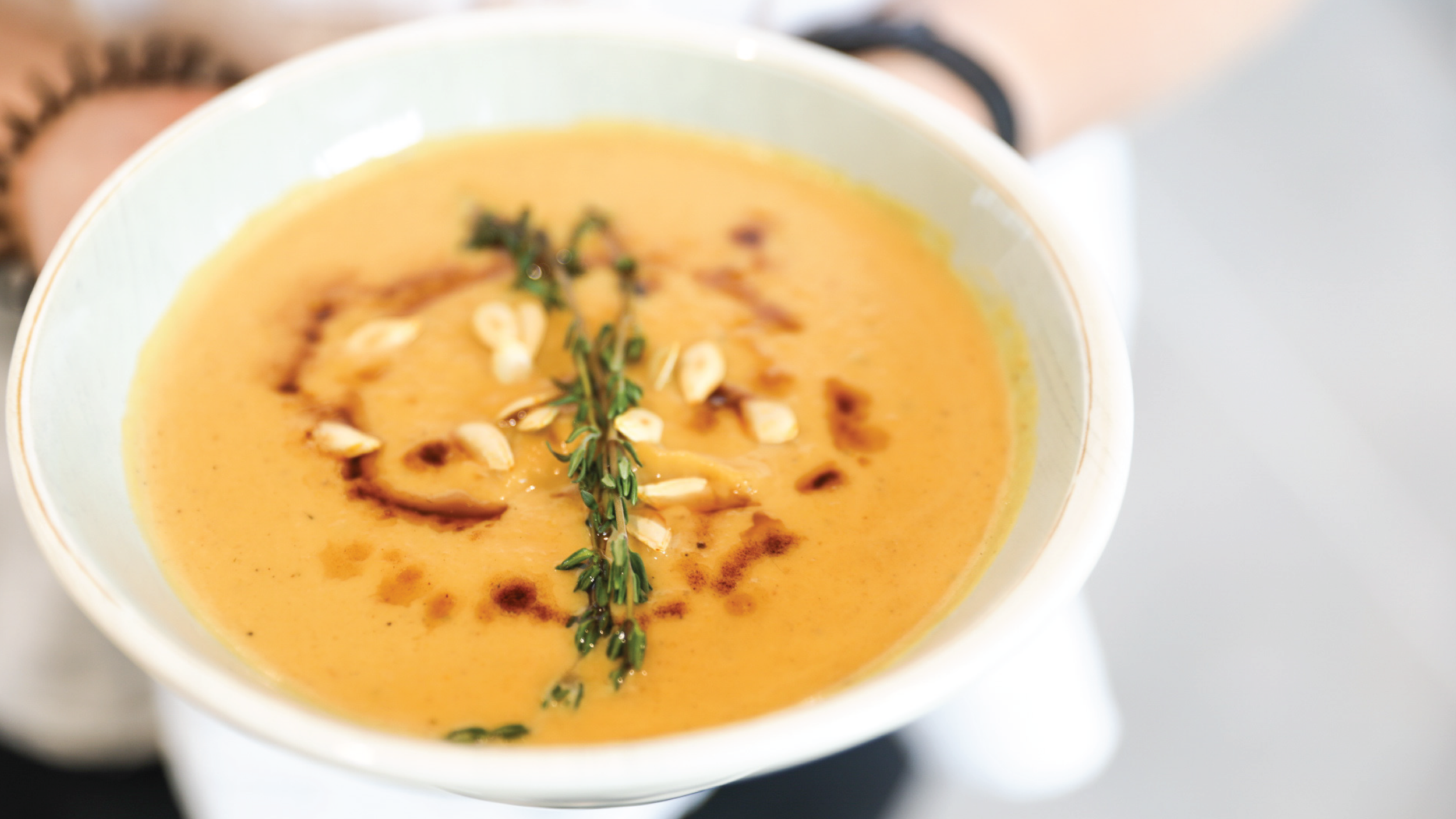 A bowl of creamy butternut squash soup garnished with a sprig of thyme, pine nuts, a drizzle of balsamic glaze, served on a white plate.