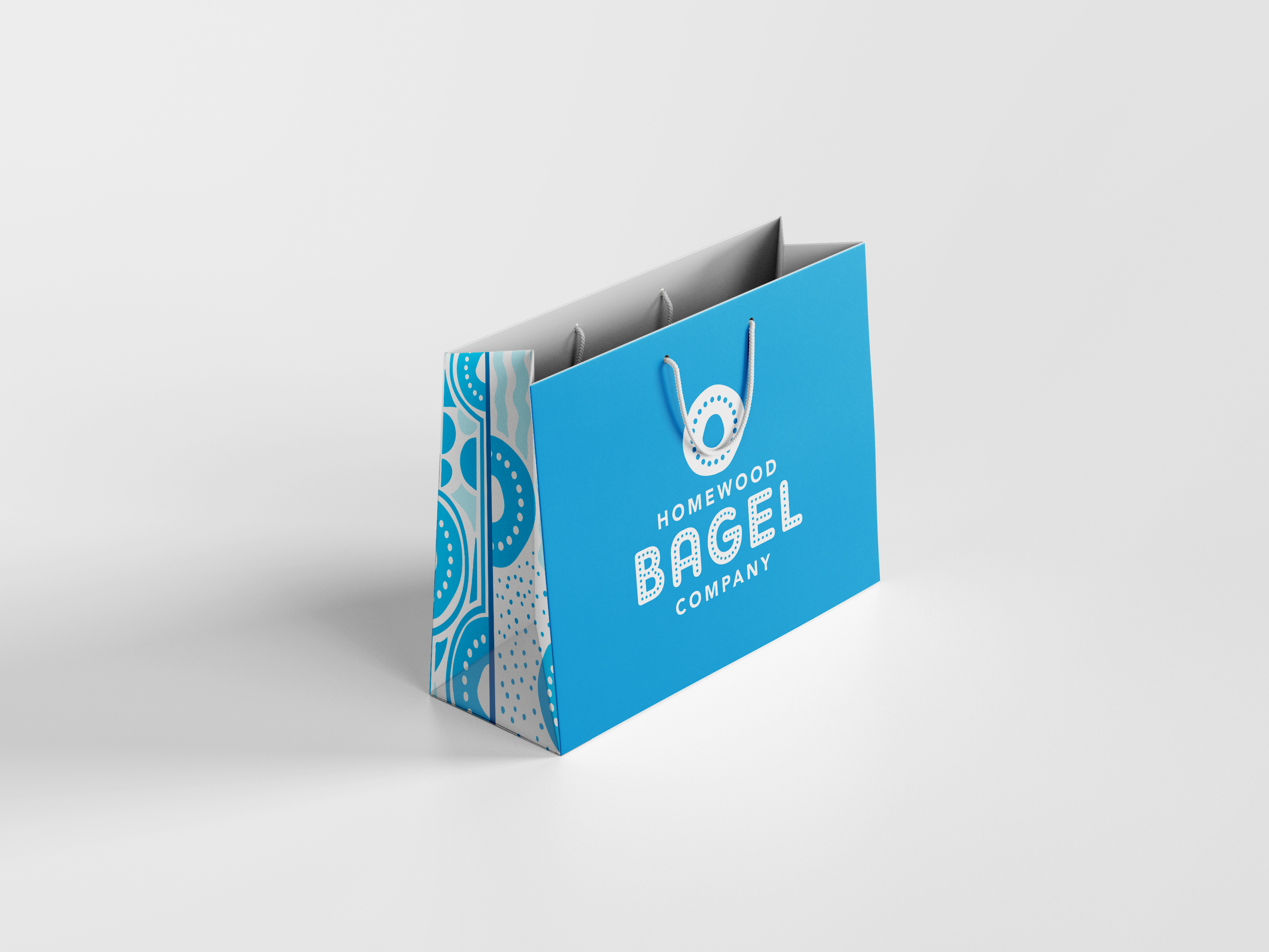 Blue shopping bag with handles, decorated with circular patterns on the side, displaying the text "Homewood Bagel Company" along with a graphic of a bagel.