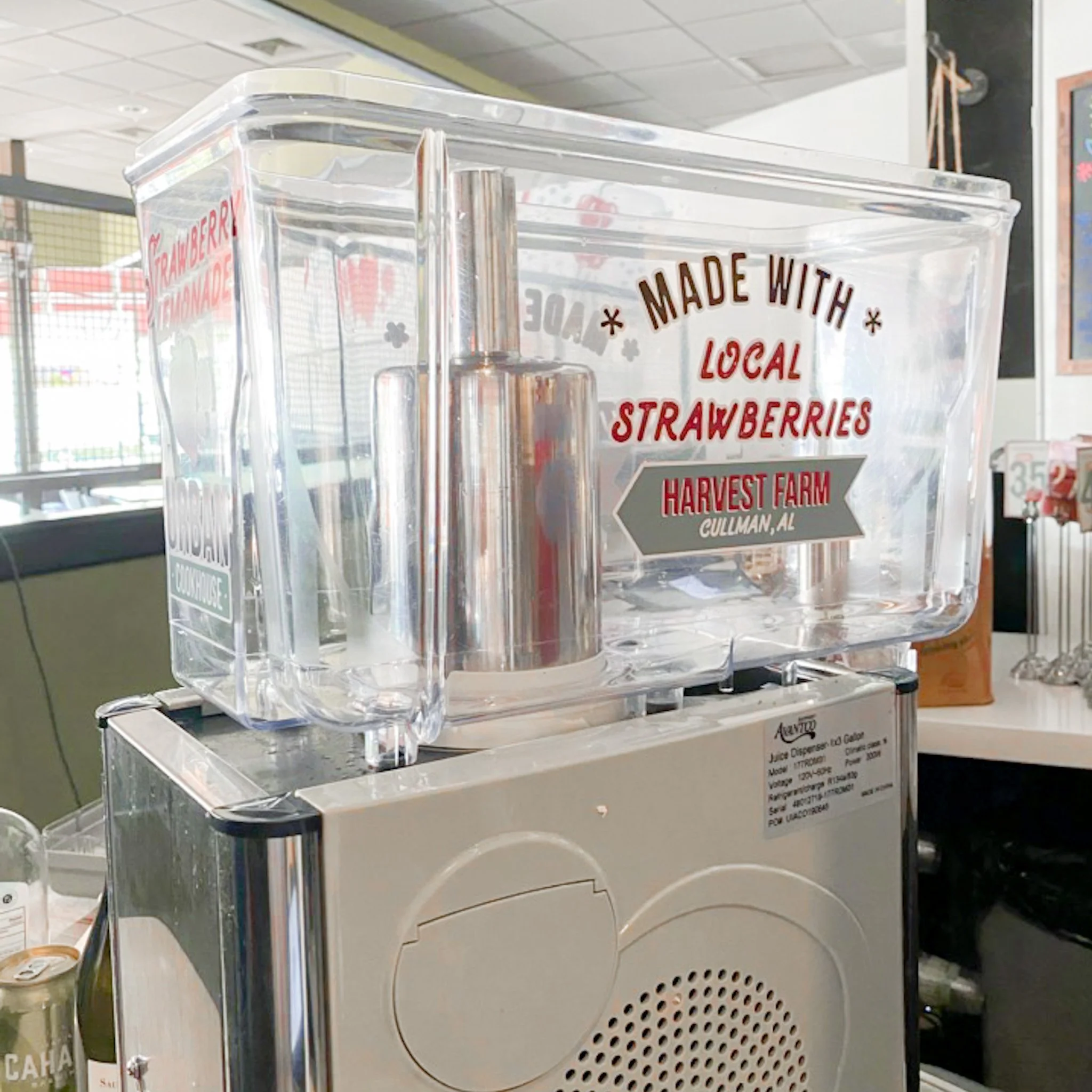 A clear juice dispenser with a label indicating it contains locally made strawberry juice from Harvest Farm in Cullman, Alabama.