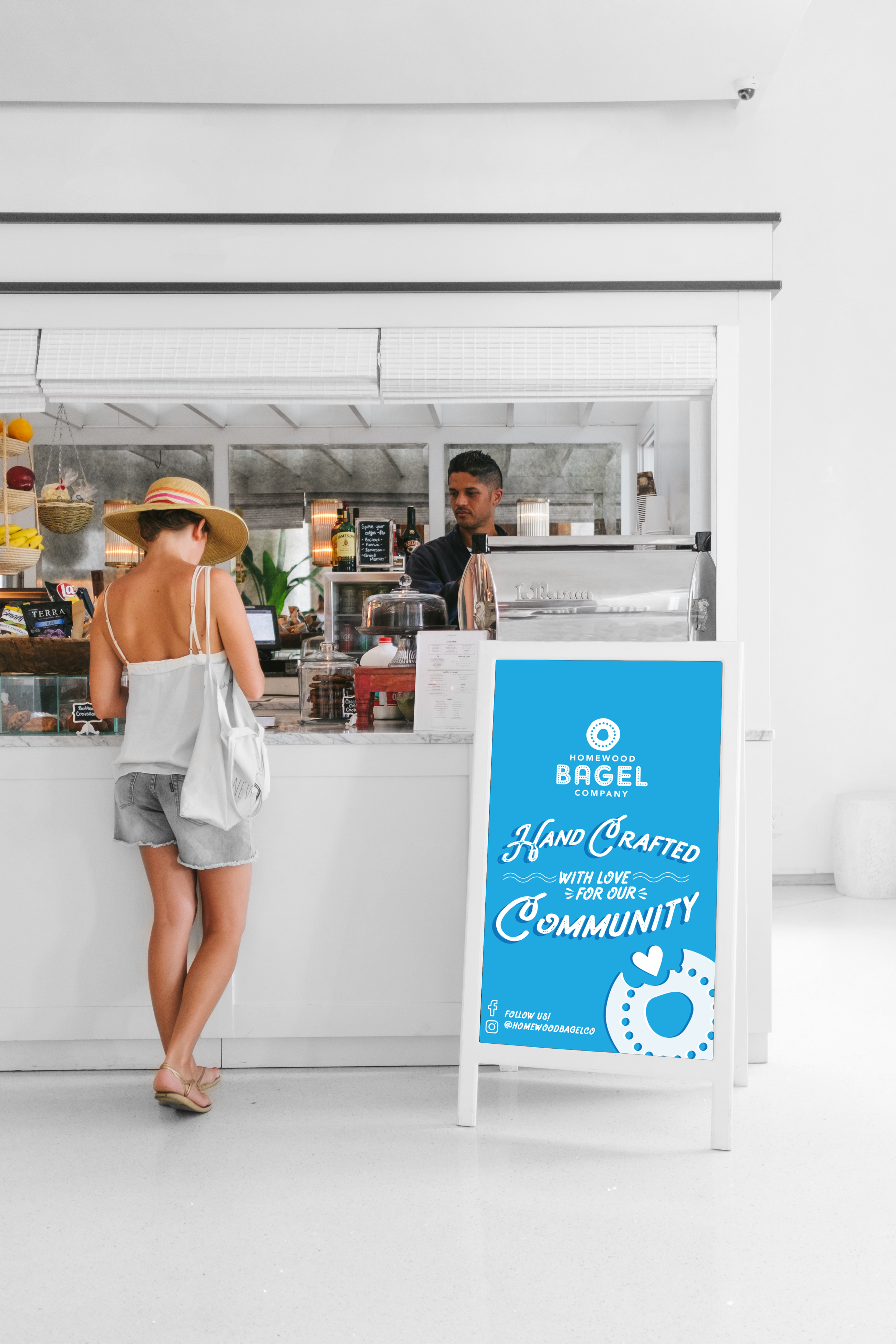 A woman orders at a cafe counter while wearing a wide-brimmed hat, white tank top, and denim shorts. A signboard outside the cafe promotes handcrafted baked goods made with love for the community.