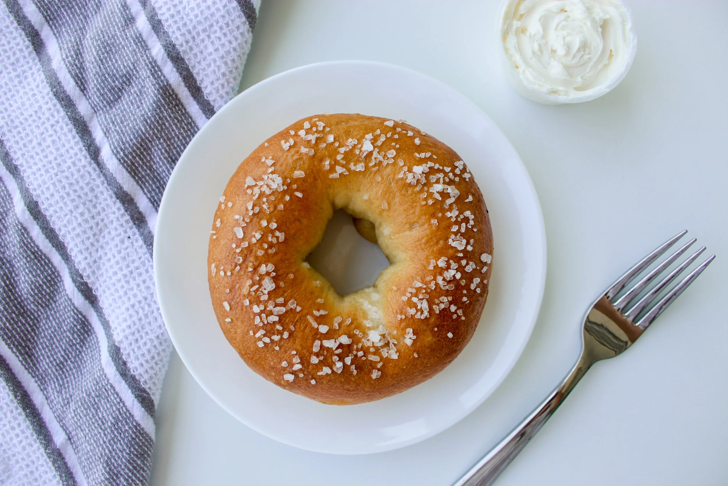 A plain bagel with coarse salt on top on a white plate, with a small bowl of cream cheese, a silver fork, and a black-and-white striped cloth napkin on a white surface.