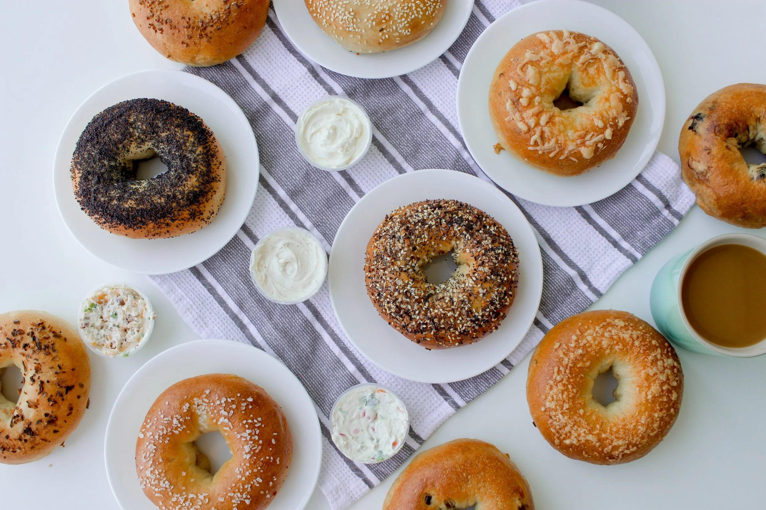 An assortment of glazed and sprinkled bagels on white plates, with small cups of cream cheese and a cup of coffee on a table with a gray and white striped cloth.