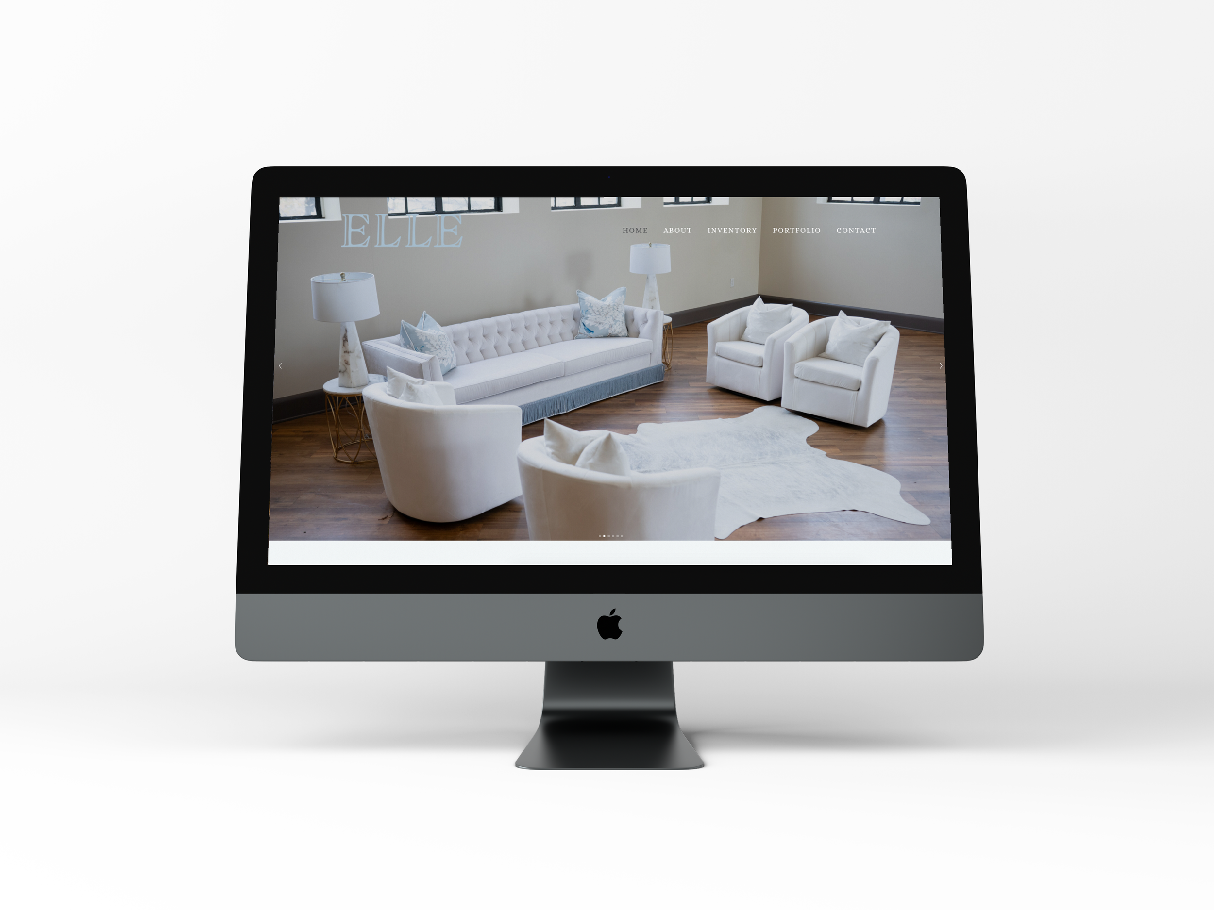 An iMac computer displaying a website with a bright, modern living room interior featuring white furniture, including a tufted sofa, two armchairs, and side tables with lamps.