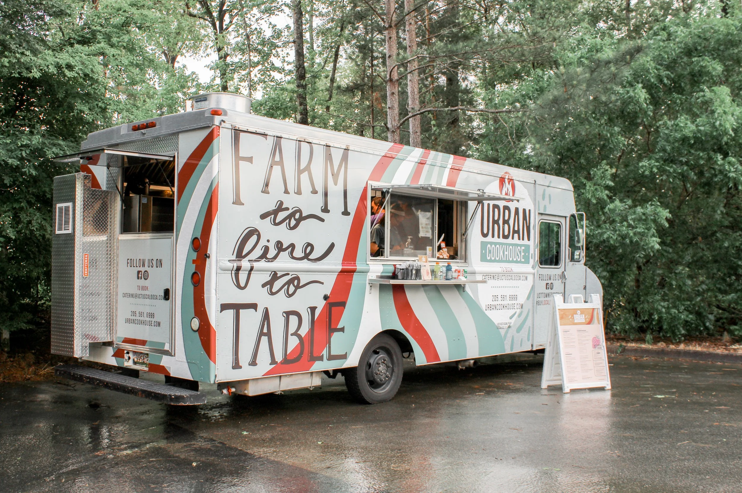 Food truck with colorful painted text and design, parked on a wet road surrounded by trees, with a menu board outside.