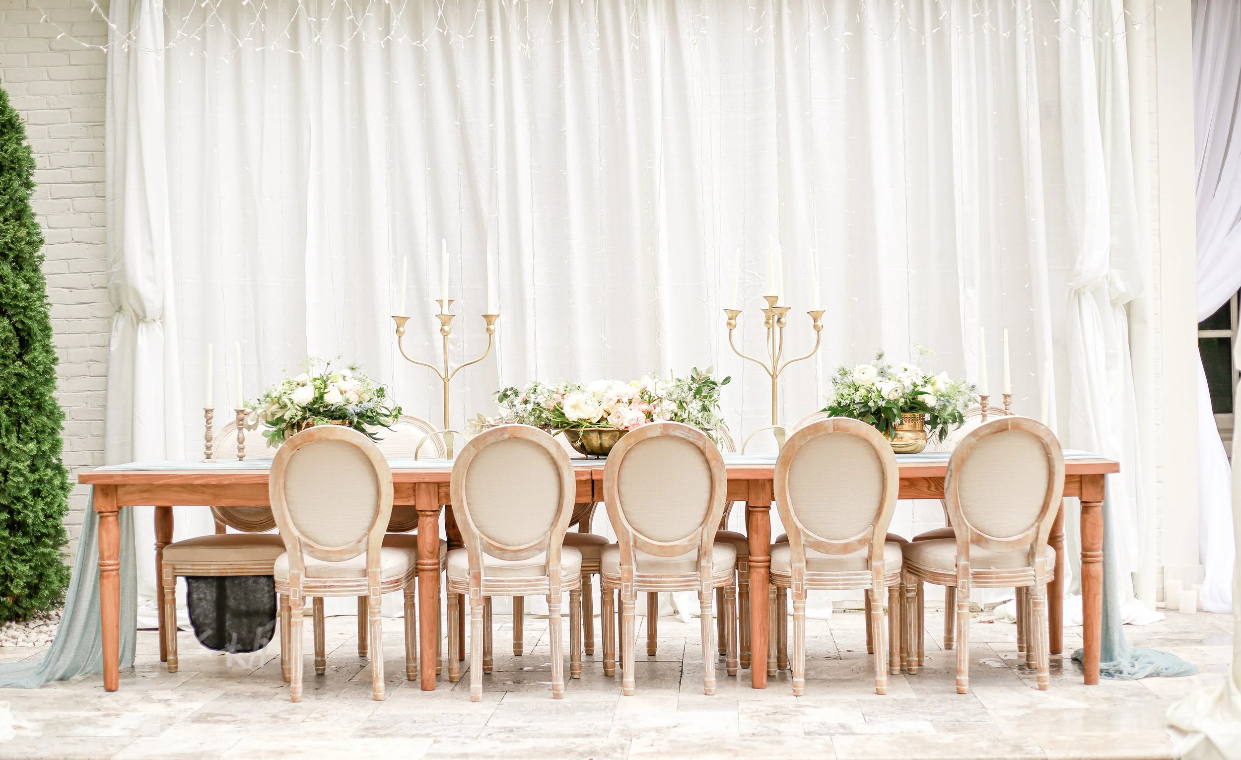 A long wooden dining table with eight upholstered chairs, set against a white curtain backdrop with floral centerpieces and gold candelabras.