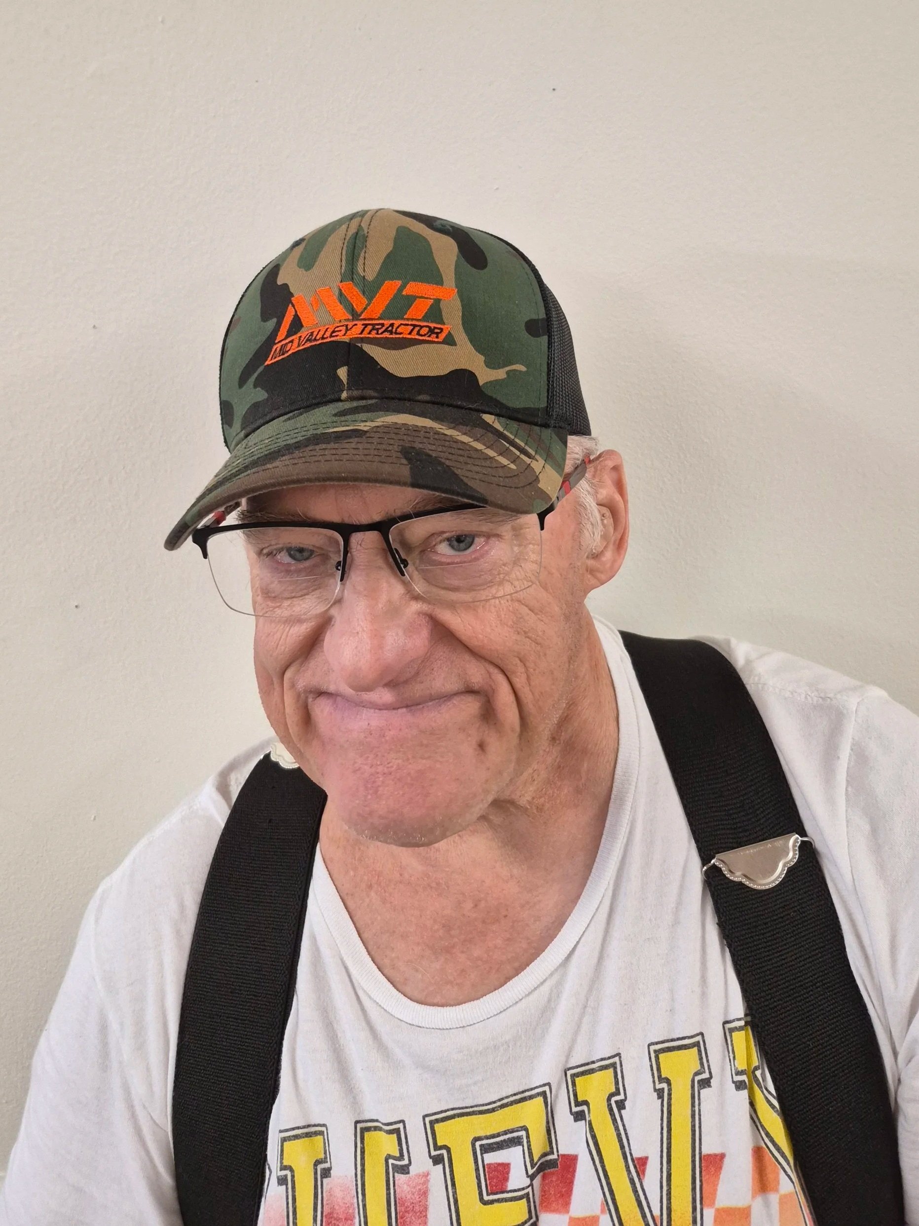 Smiling man in glasses wearing a Black Panther T-shirt and a cap, sitting in front of framed documents and plants.