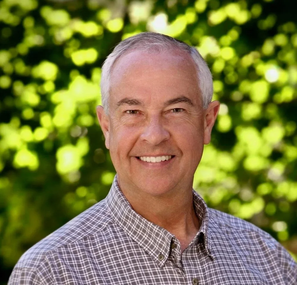 A smiling middle-aged man with short gray hair wearing a checkered shirt, standing outdoors with green foliage in the background.